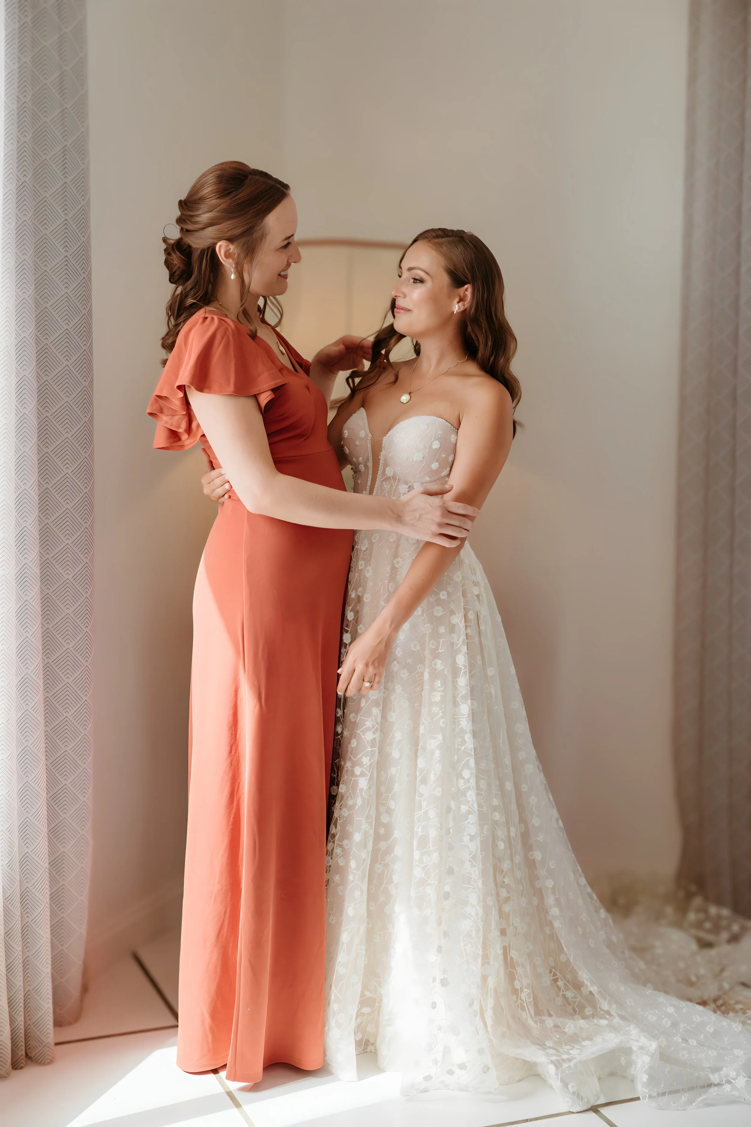 A bride in a strapless wedding gown embraces a woman in an orange dress, both smiling and looking at each other inside a warmly lit room.