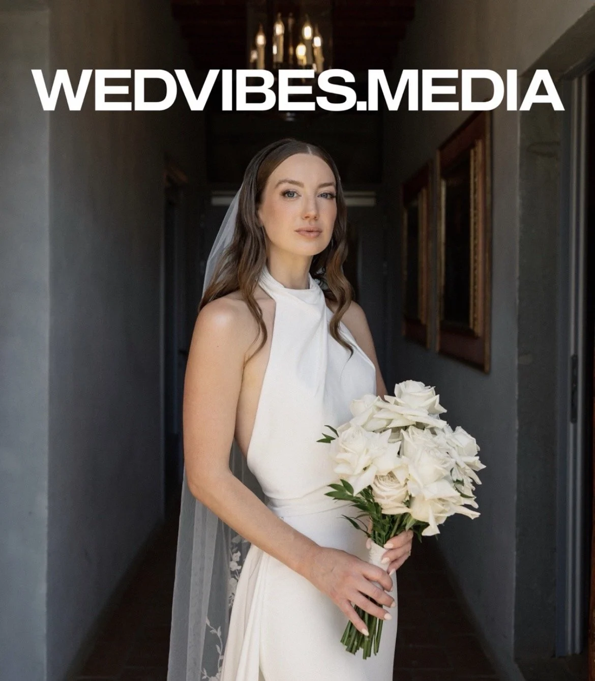 A bride with long brown hair in a white wedding dress holding a bouquet of white roses standing in a dimly lit hallway with framed pictures on the wall.
