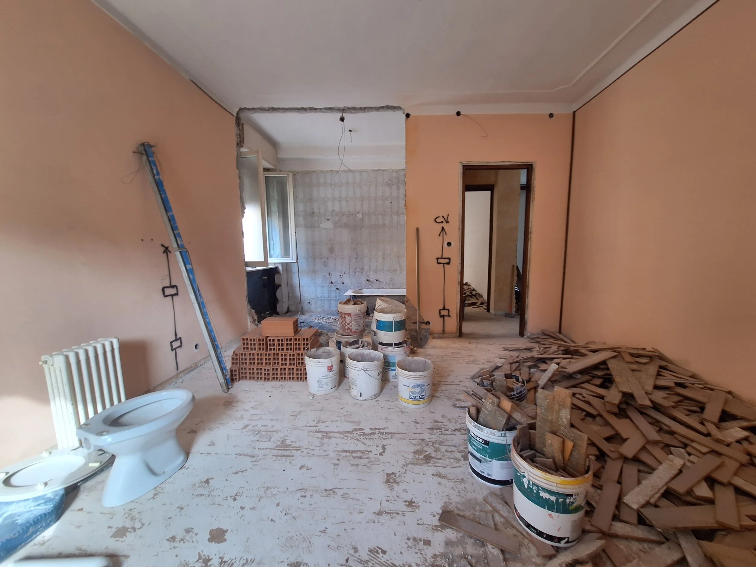 Interno di una stanza in ristrutturazione con pareti dipinte di color pesca. Sul pavimento ci sono mattoni, tubi e secchi di vernice. Un'area sembra essere in fase di demolizione, con un'apertura in una parete e pareti di sottofondo sfoggiate. Un ter