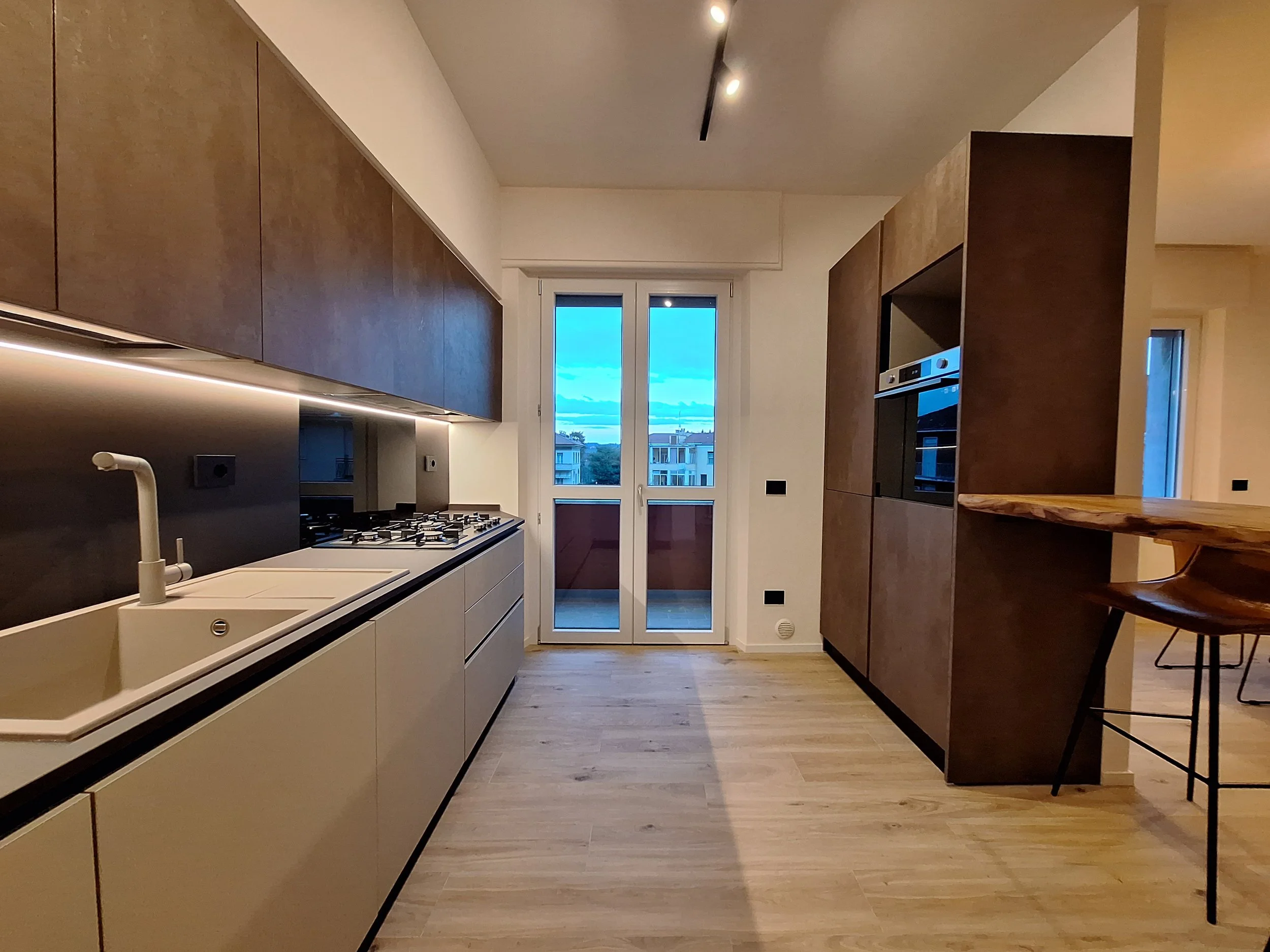 Cucina moderna con lavello bianco, piano cottura a gas, armadietti beige superiori e inferiori, grande finestra con vista sulla città, pareti bianche, pavimento in legno chiaro, schienale nero.