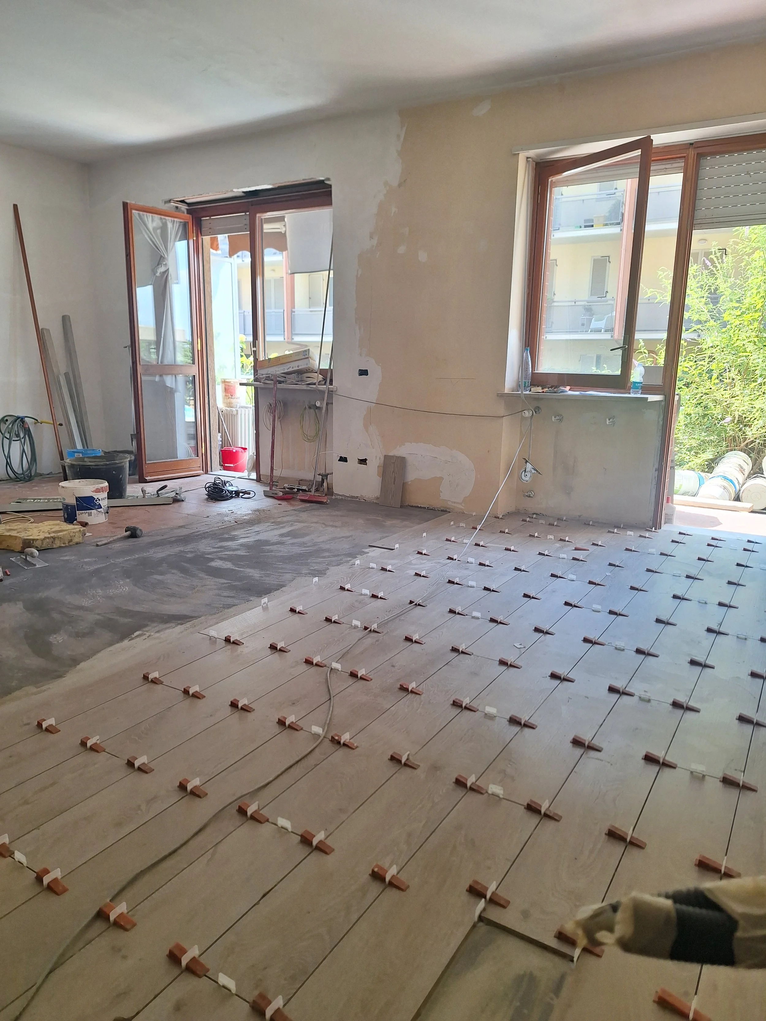Interno di una casa in ristrutturazione con pavimentazione in installazione, grandi finestre aperte, alcuni strumenti e materiali da costruzione.