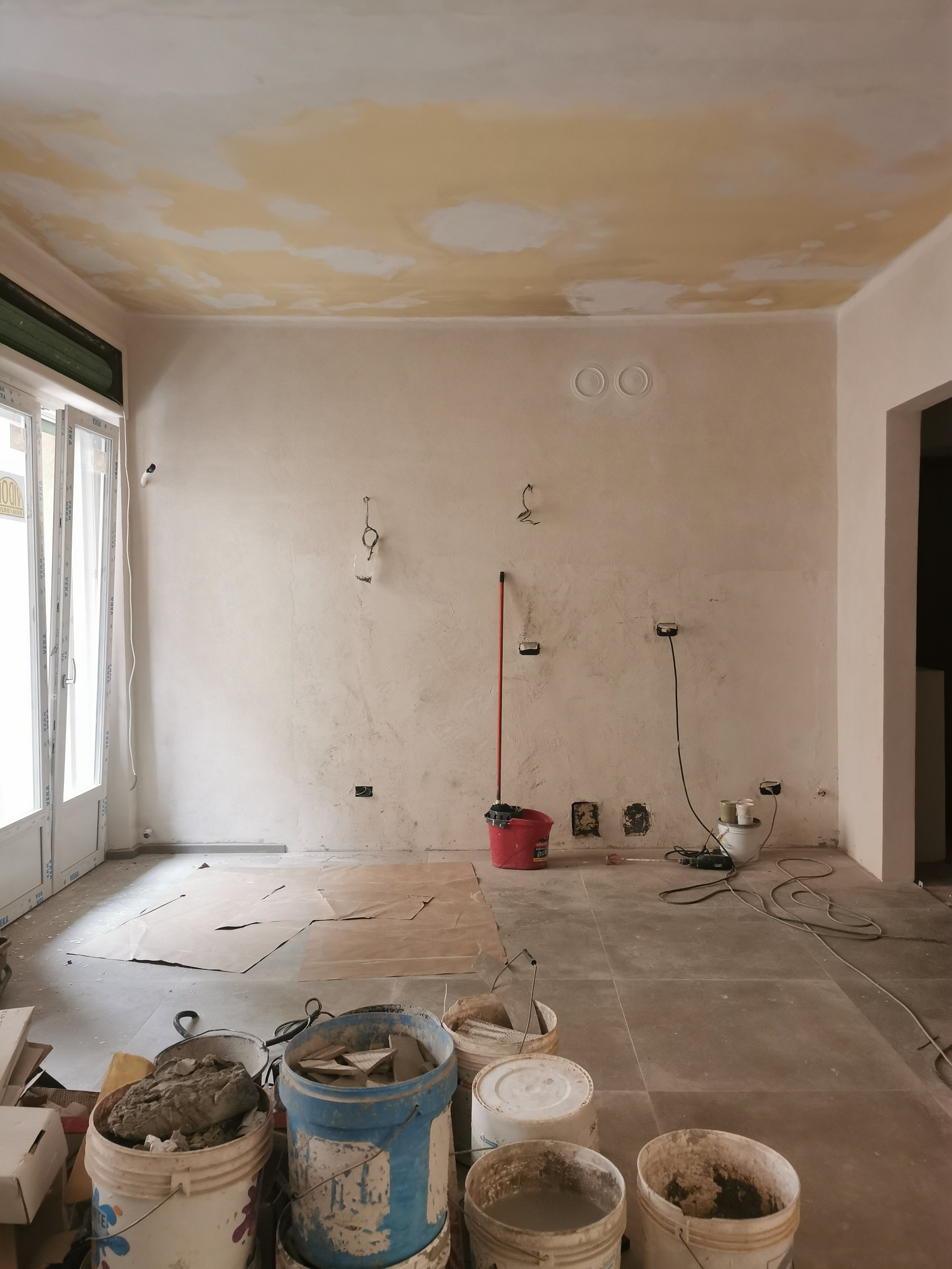 Interno di una stanza in fase di ristrutturazione con pareti e soffitto in corso di tinteggiatura, attrezzi e contenitori di materiali edili sul pavimento, e finestre sul lato sinistro.