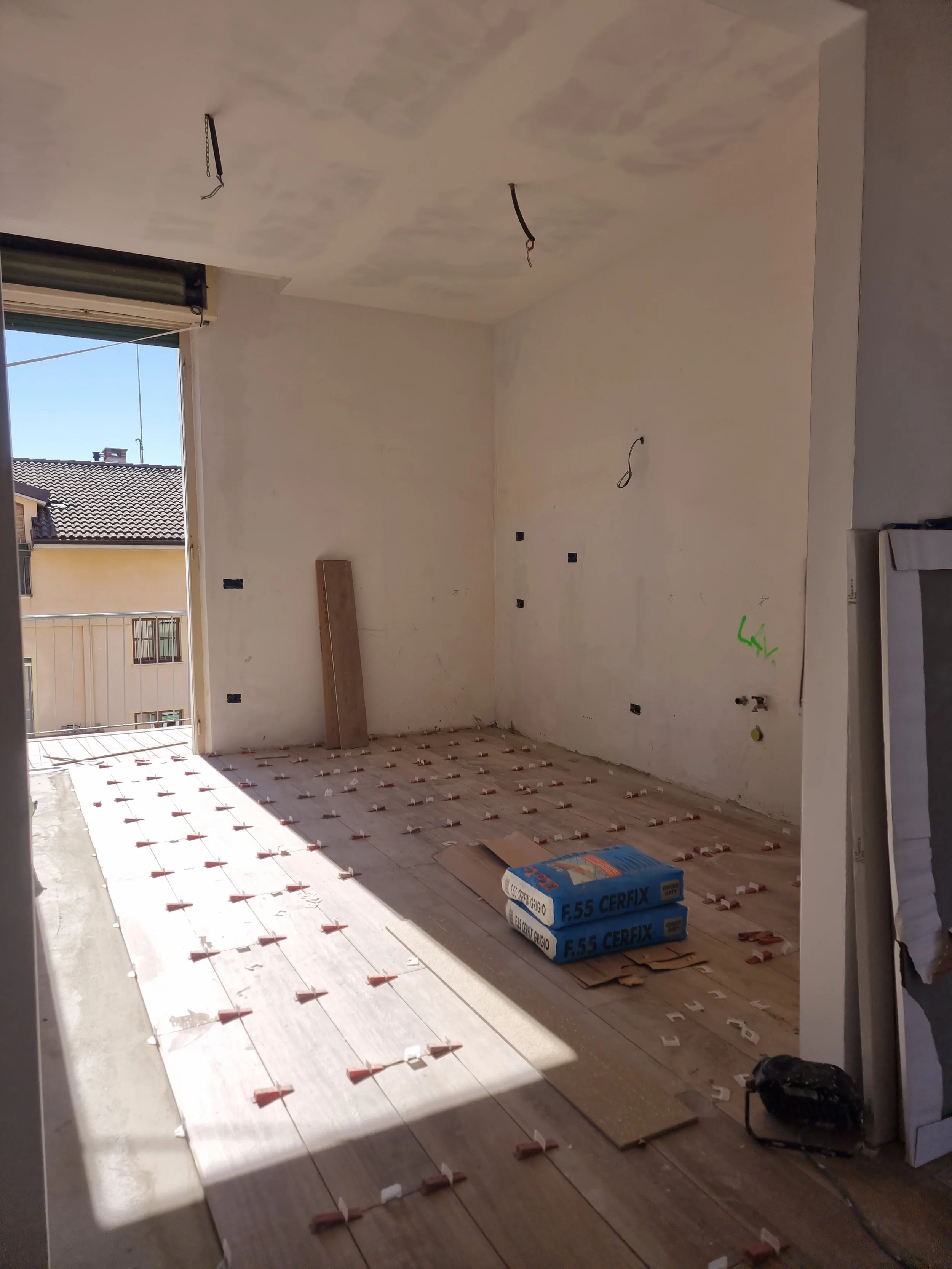 Stanza in costruzione con pavimentazione in fase di posa, luci sospese, pareti non finite, alcune scatole di materiali edili.