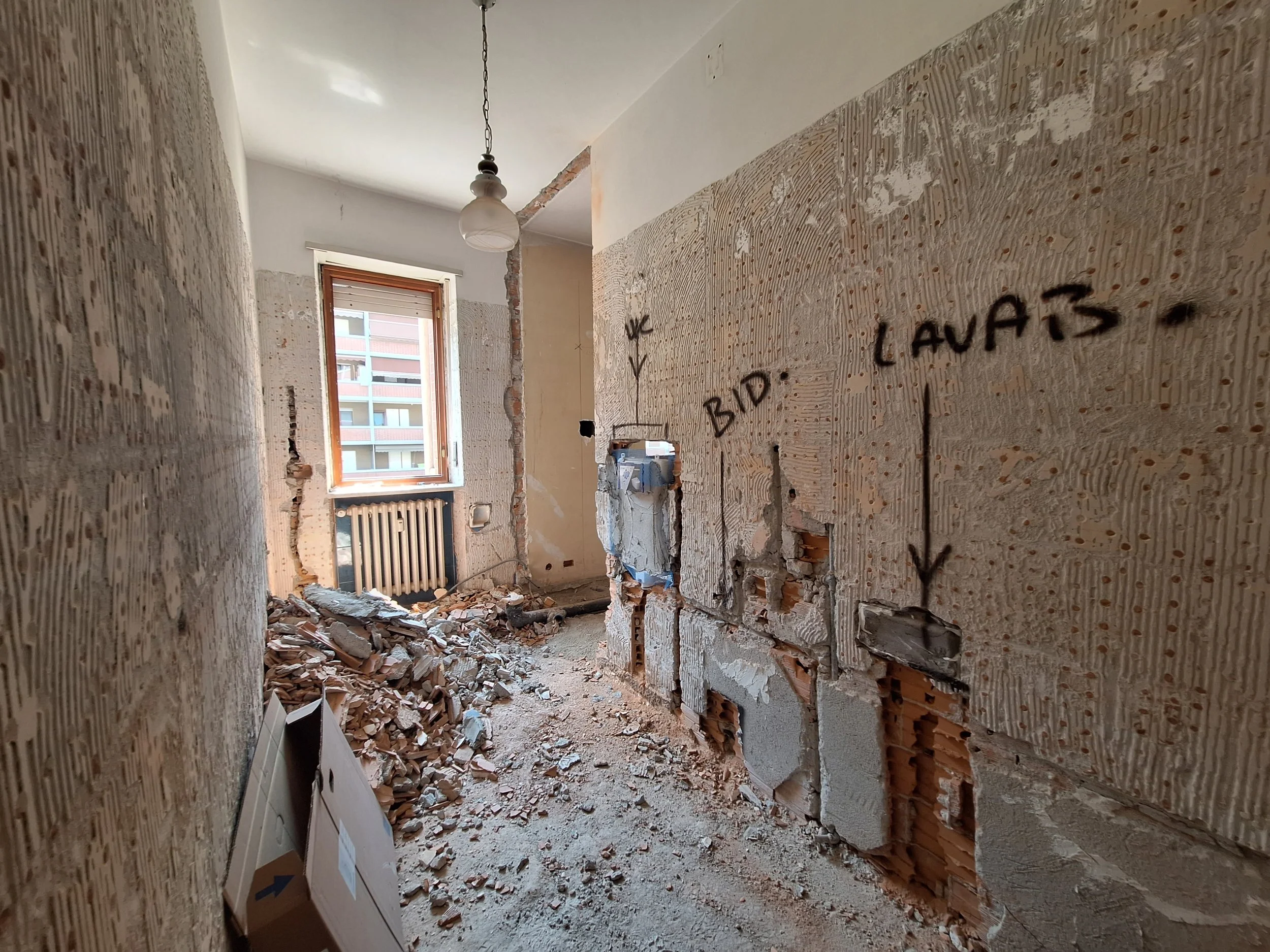 Stanza in ristrutturazione con pareti danneggiate, pavimento coperto di detriti e una finestra