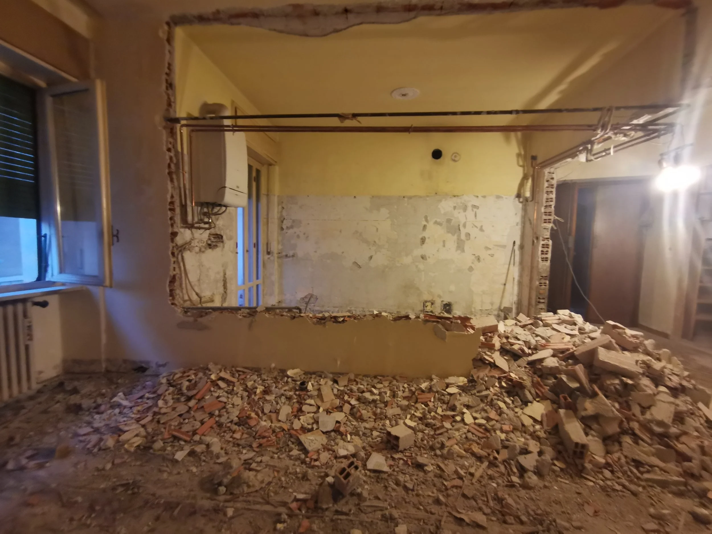 Stanza in fase di ristrutturazione con muri in demolizione, mattoni e polvere sul pavimento, finestra e porta aperte, e tubi sospesi dal soffitto.