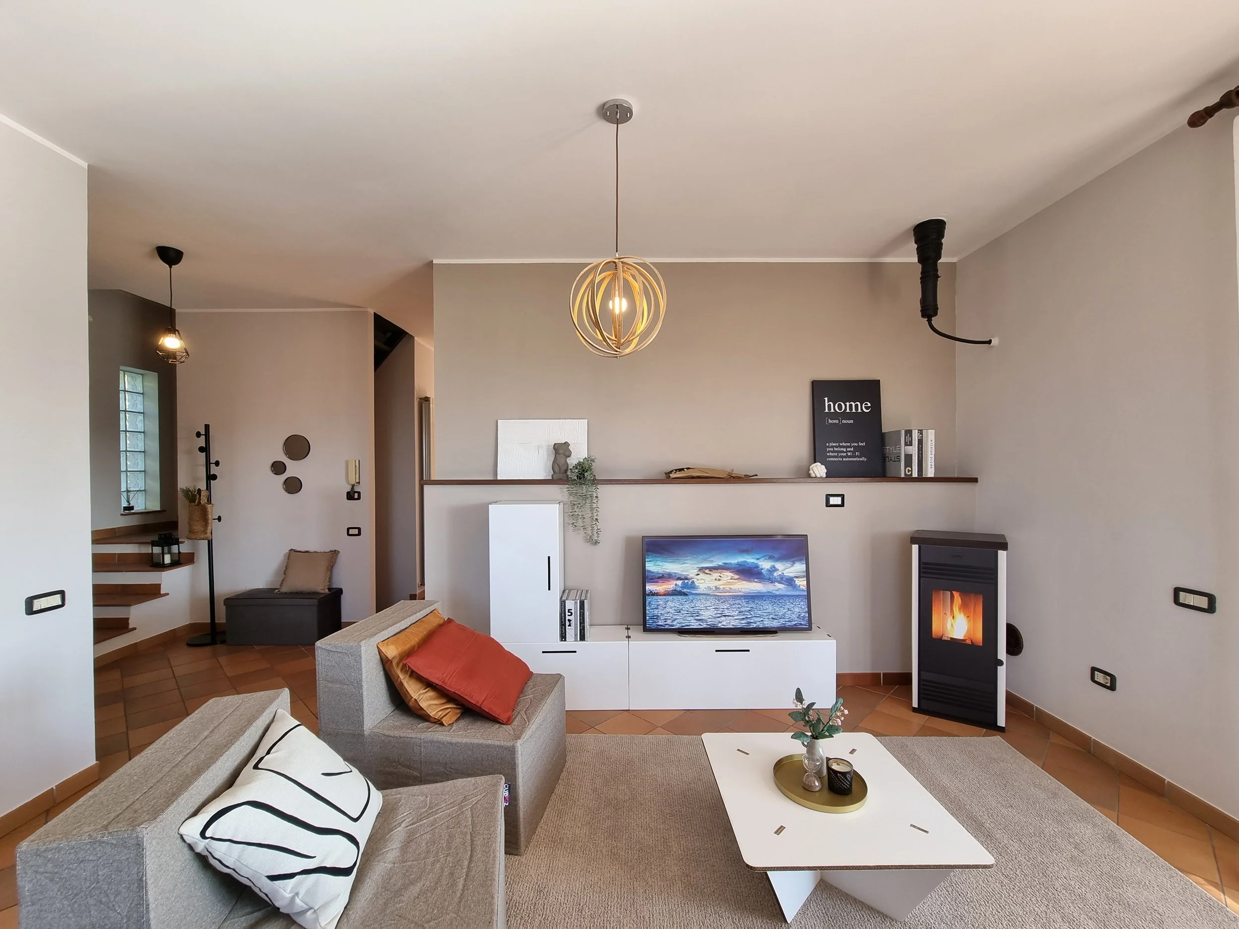 Soggiorno moderno con divano, tavolino, televisore, camino a pellet, decorazioni e scaffale con libri, illuminazione sospesa, pareti chiare e pavimento in ceramica