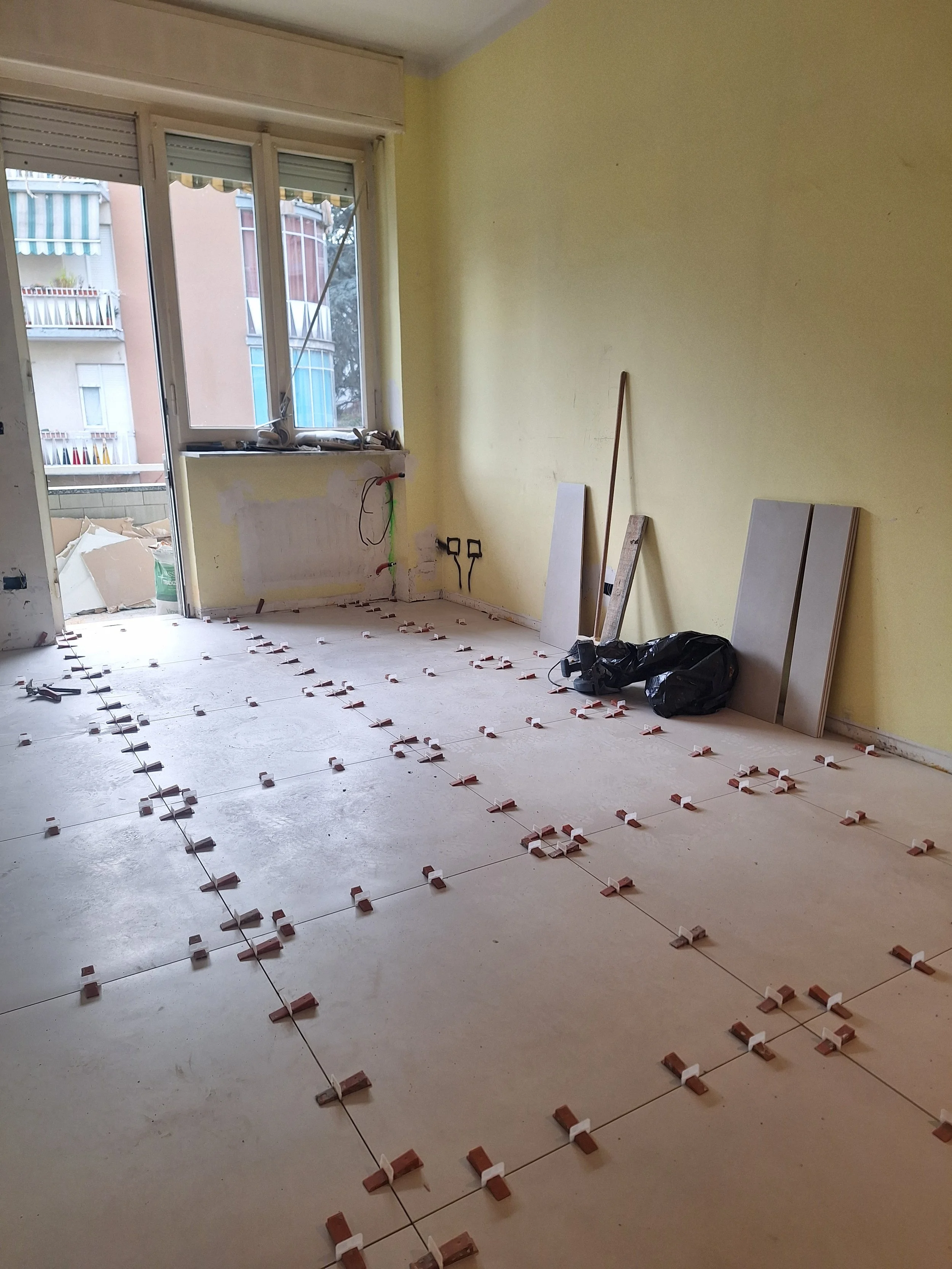 Stanza in fase di ristrutturazione con piastrelle non ancora posate, strumenti e materiali di lavoro, finestra aperta con vista sui palazzi vicini.
