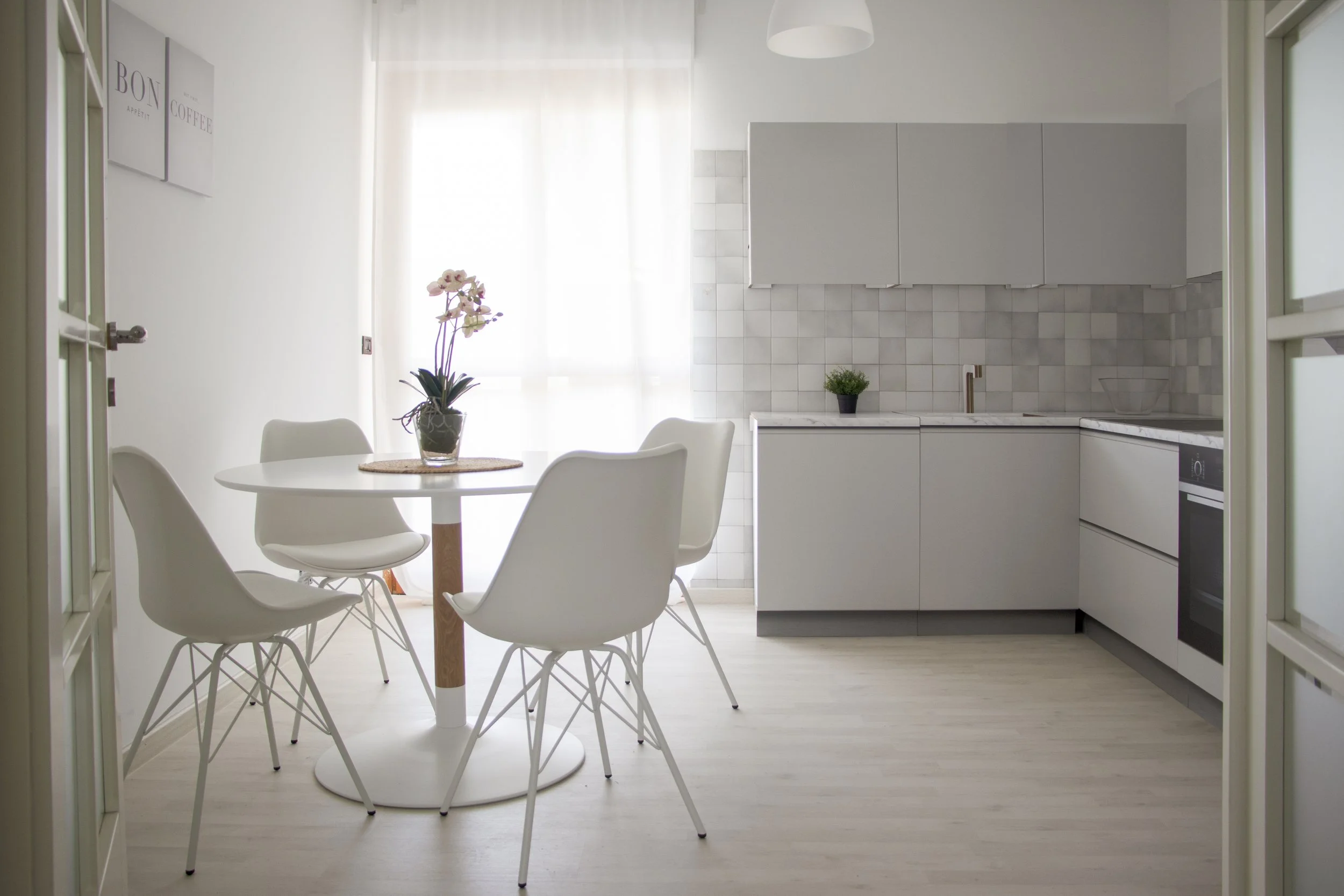 Interno di una cucina luminosa con tavolo rotondo bianco, quattro sedie bianche e un vaso di fiori sulla tavola. Cucina moderna con armadi grigi, piastrelle chiare e due piante, una sul tavolo e una sul lavello.
