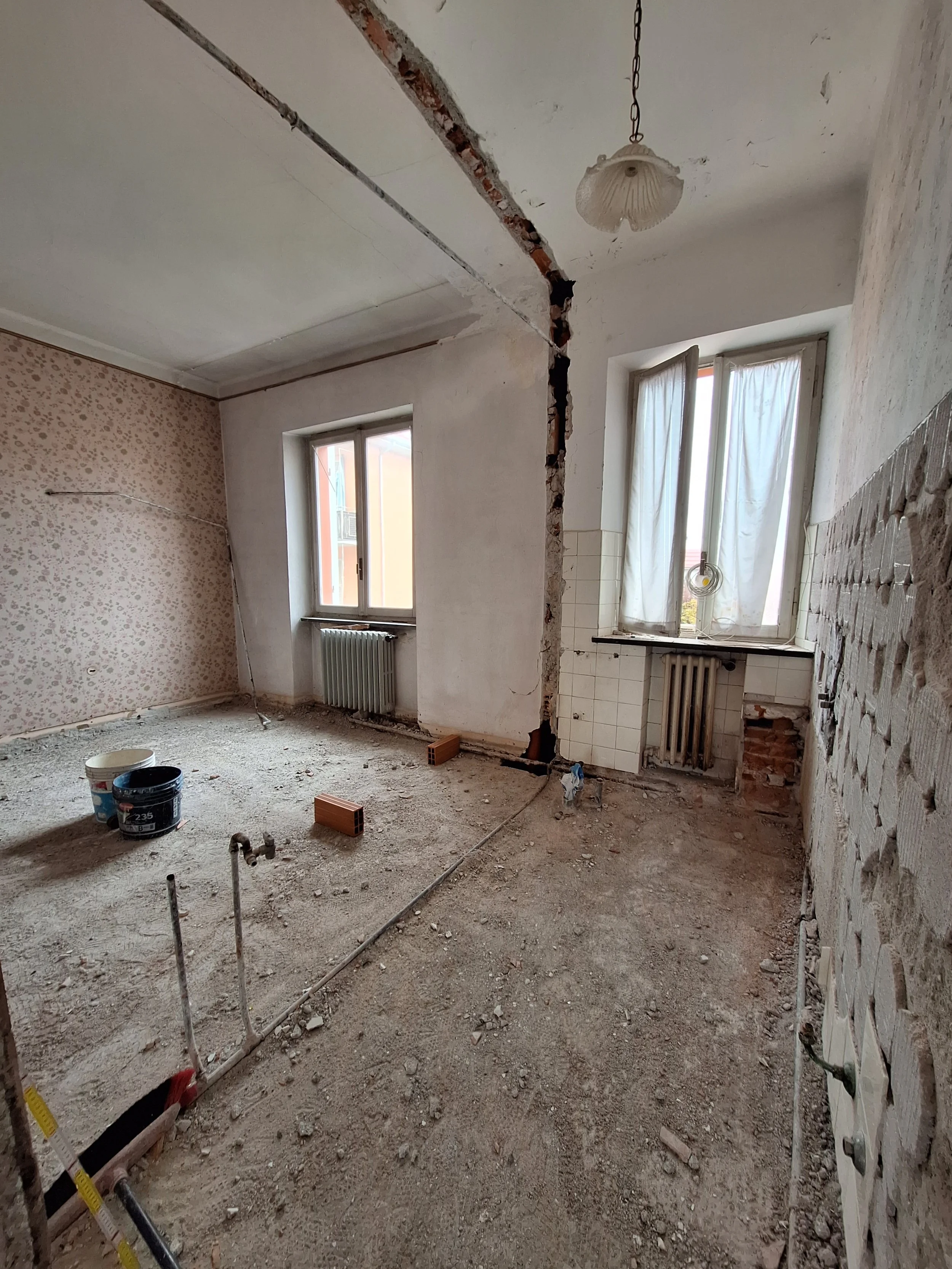 Interno di una stanza in ristrutturazione con pavimento di cemento, pareti in parte smantellate, due finestre con tende bianche, alcuni mattoni e tubi esposti, utensili e materiali da costruzione sul pavimento.