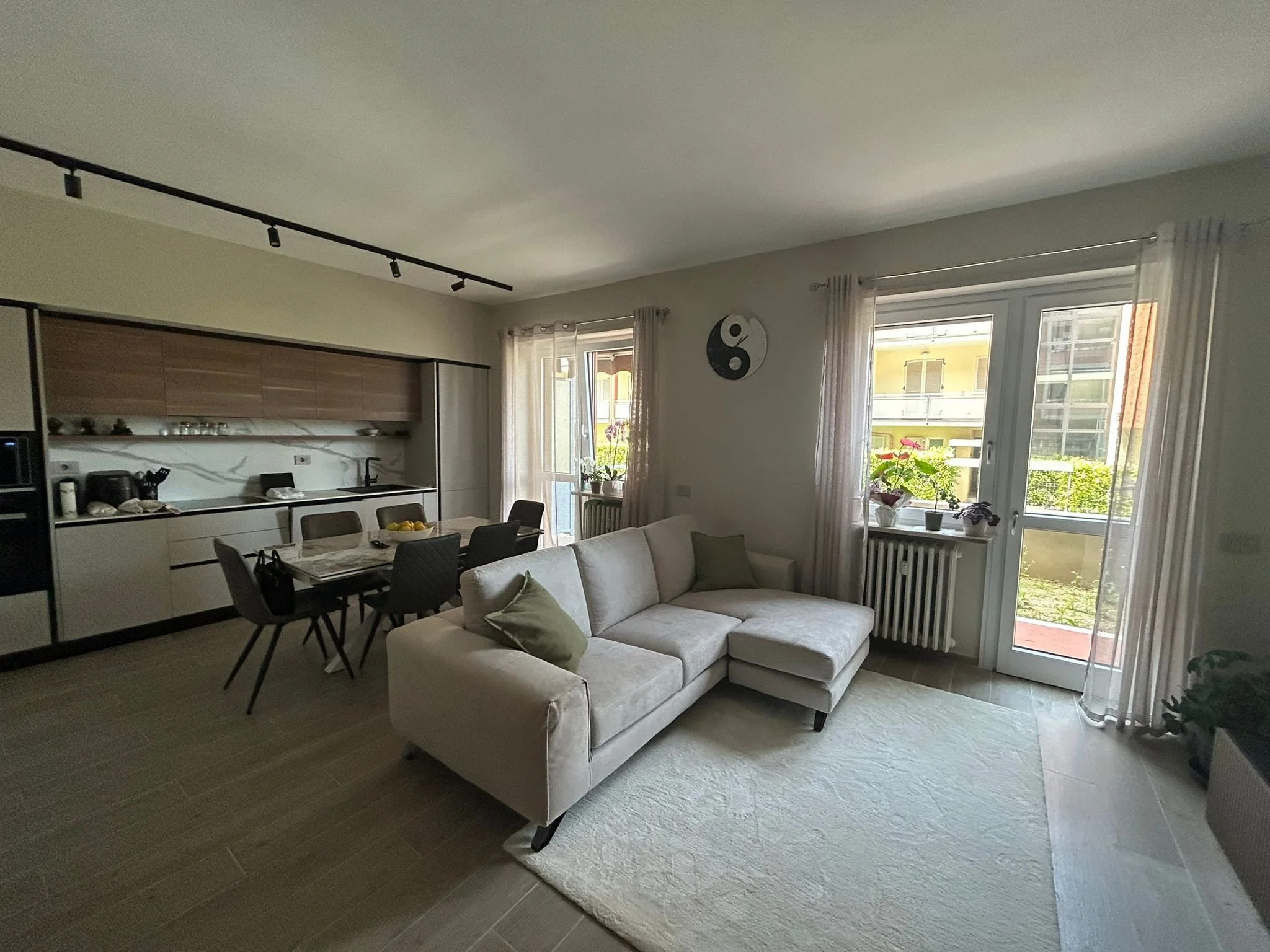 Interno di un soggiorno con divano bianco, tavolo da pranzo nero con sei sedie, cucina moderna sullo sfondo, finestre con tende leggere, decorazioni minimaliste e piante sul davanzale.