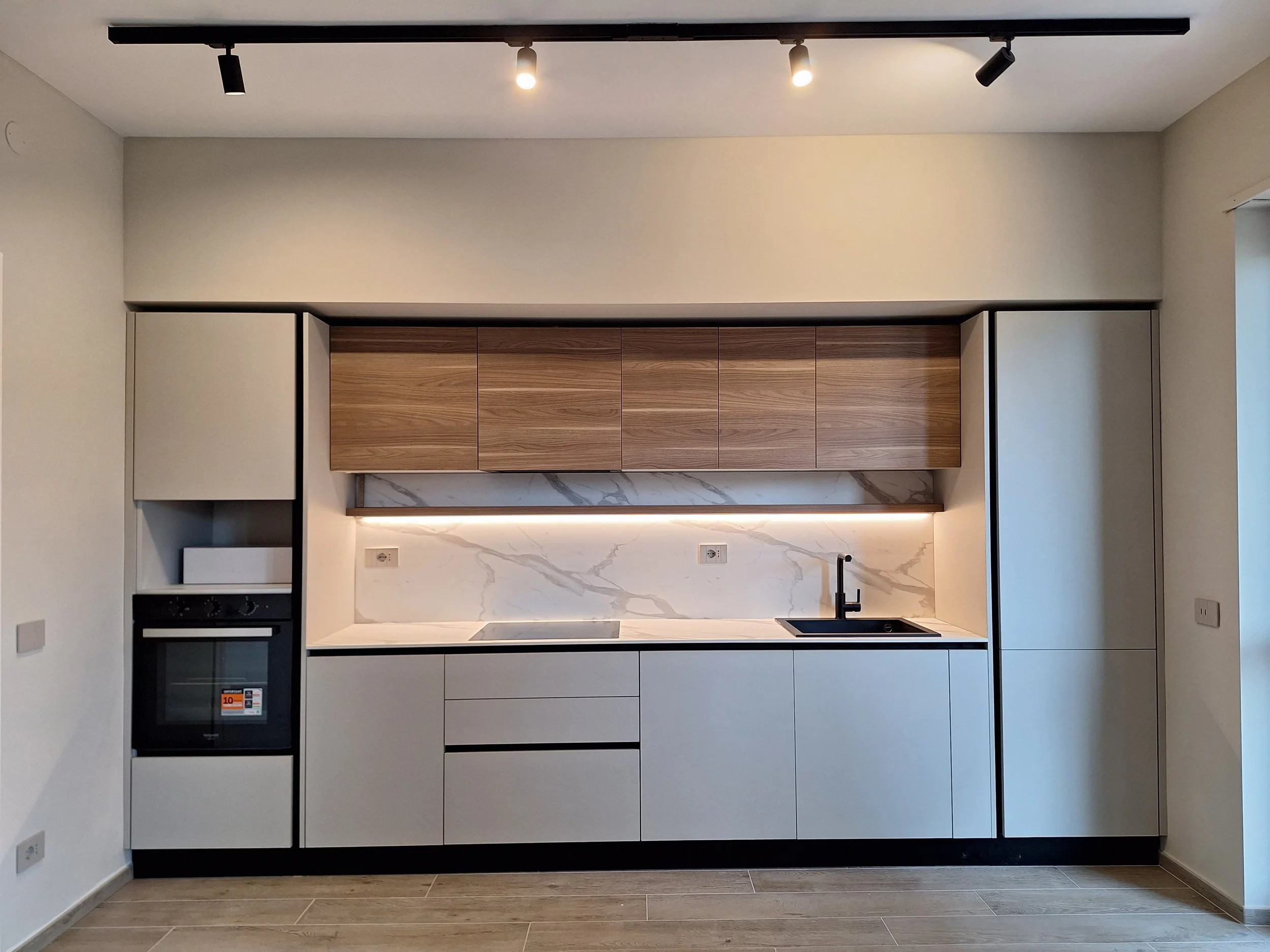 Cucina moderna con ante bianche, pensili in legno e parete in marmo, lavabo nero e piano cottura.