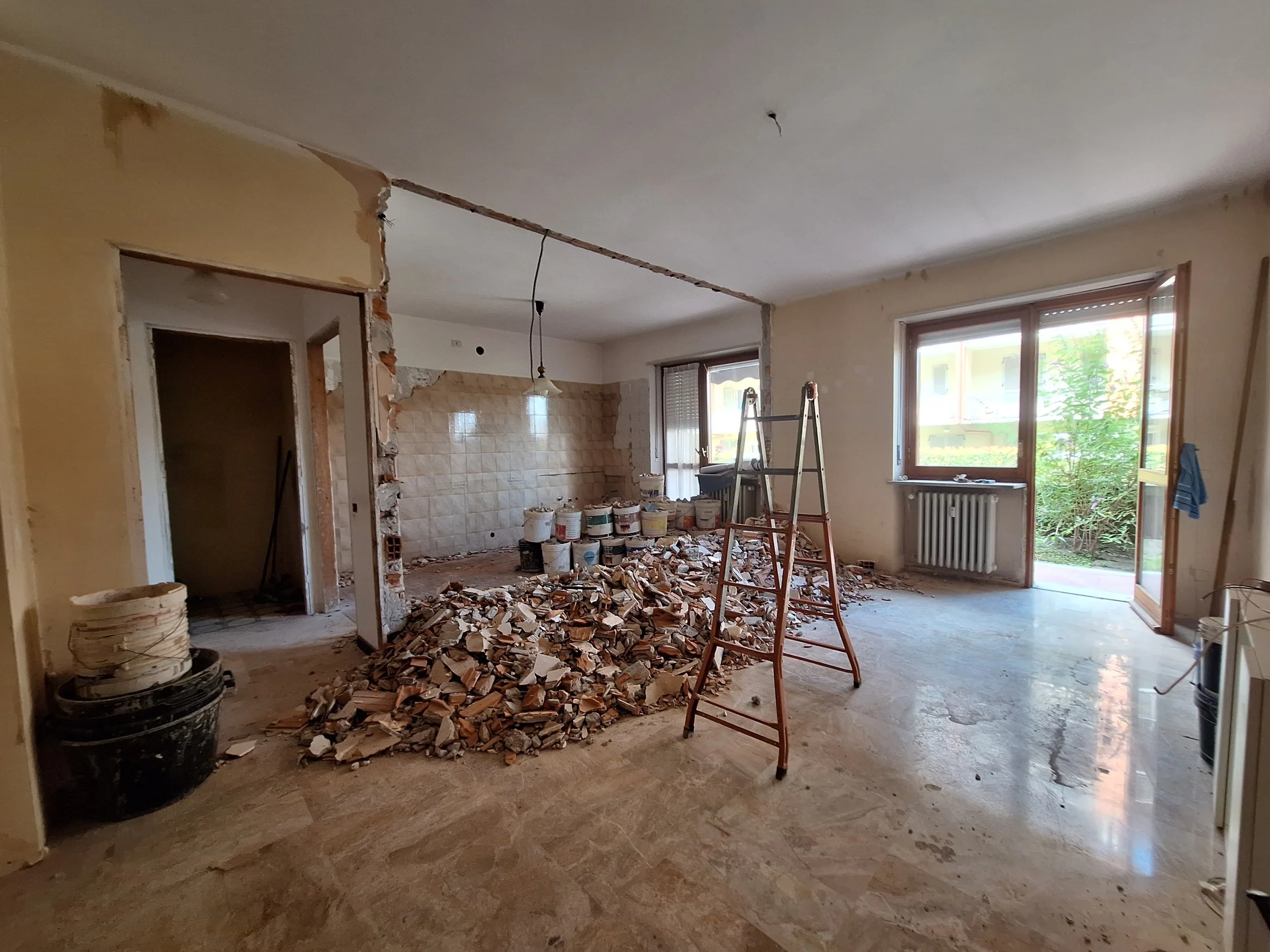 Interno di una casa in ristrutturazione con muro abbattuto, detriti e scale inizia di posa, vasi di vernice e finestre che si affacciano su un giardino verde.