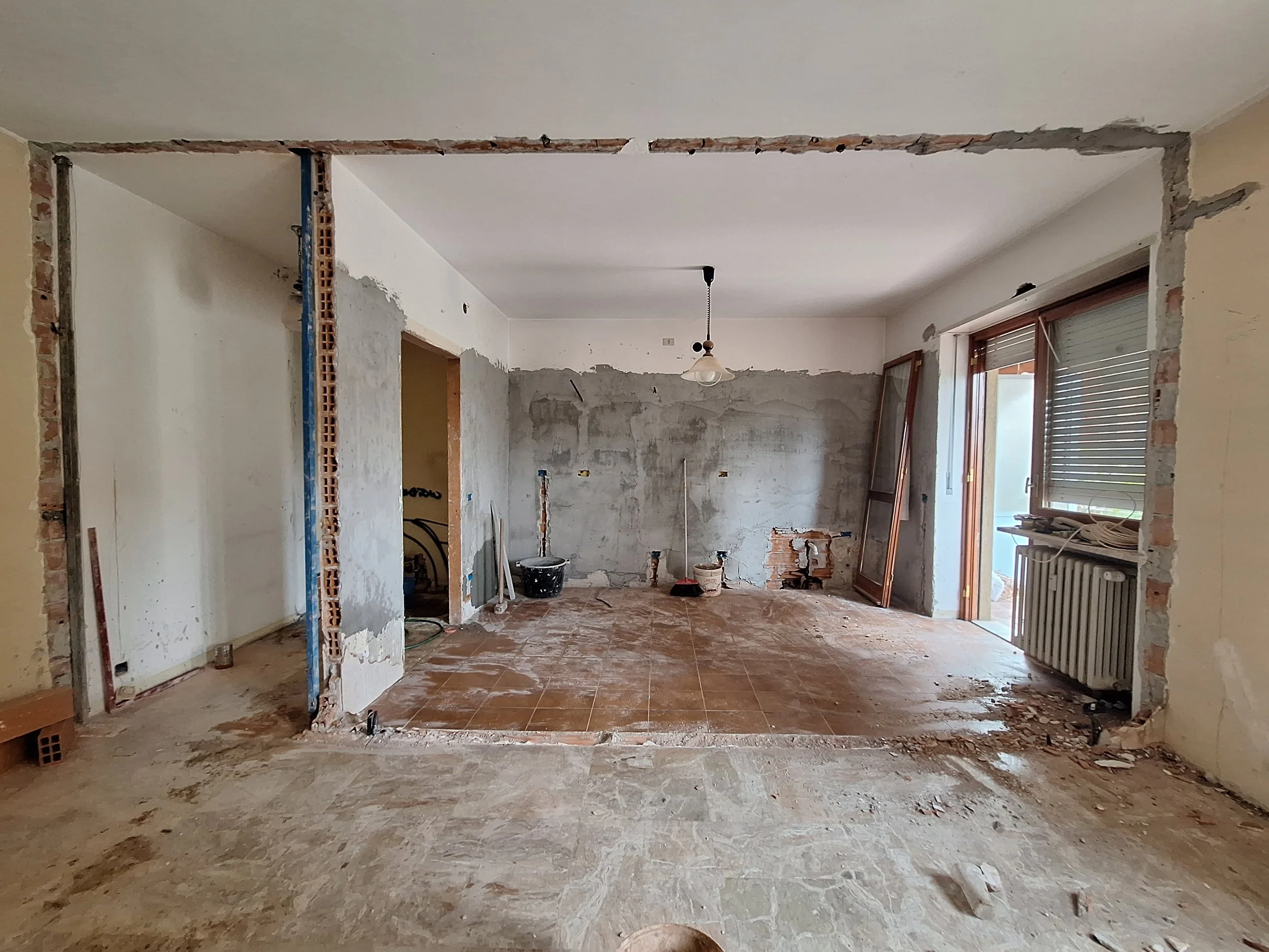 Interno di una casa in ristrutturazione con pareti in fase di scasso, senza pavimento e con alcune finestre e porta che portano a un balcone.
