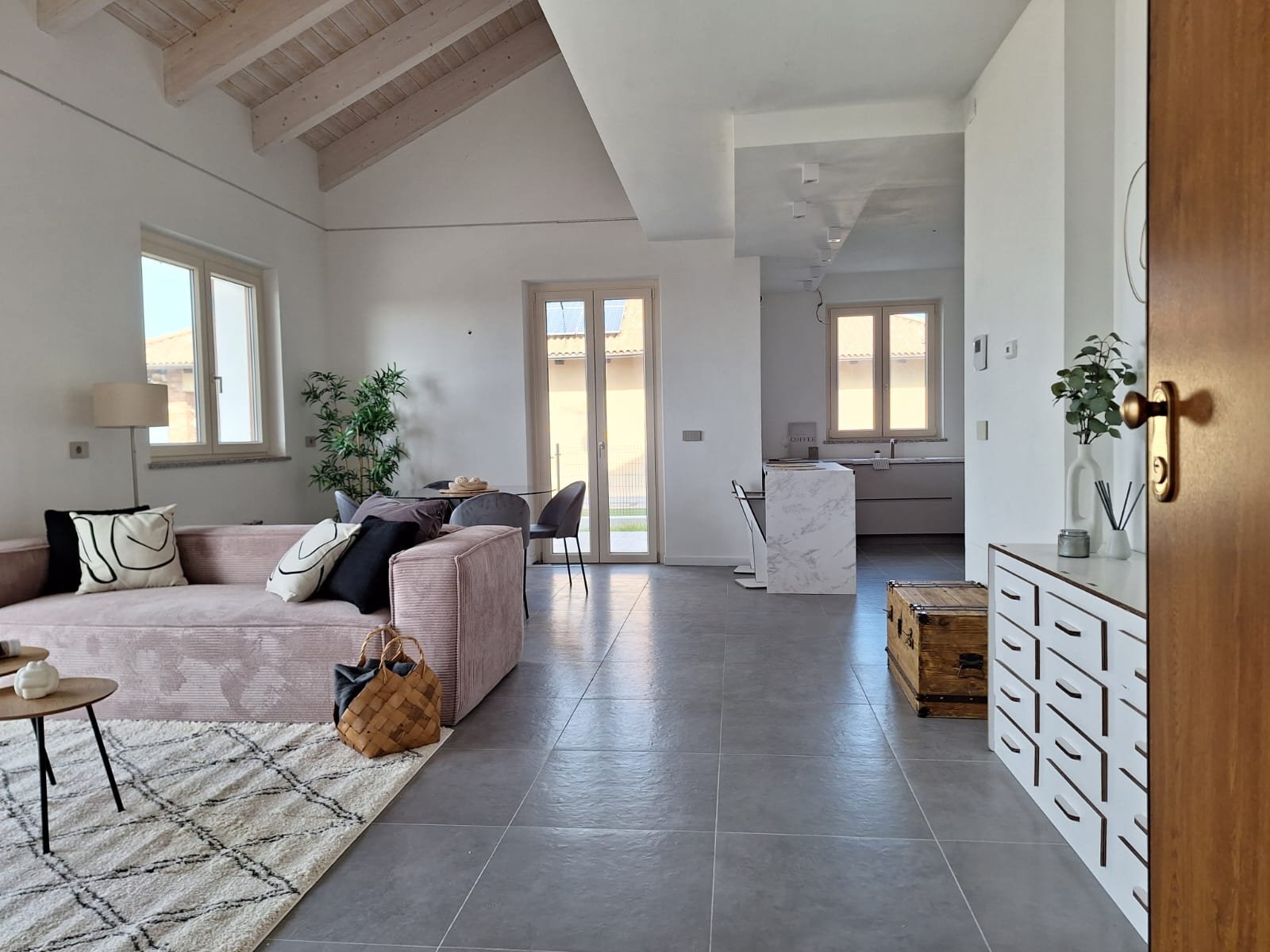 Interno di un soggiorno moderno con soffitto in legno, divano rosa, sedie nere, tavolo in vetro, grandi finestre, piante, e un'area cucina con piano in marmo.