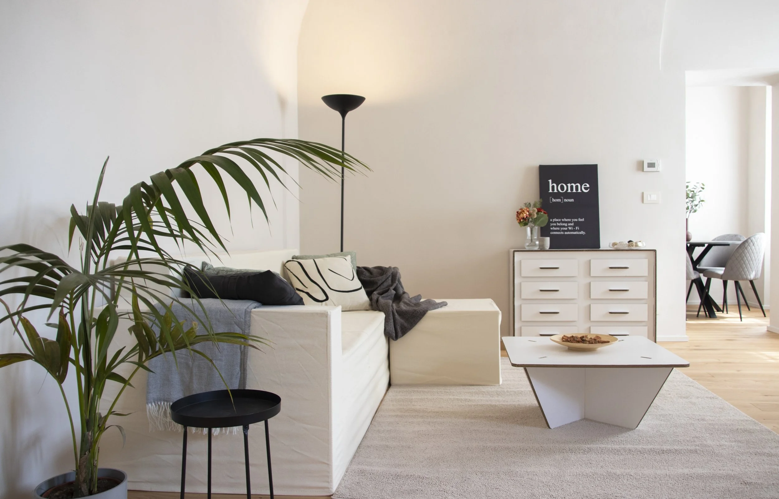 Soggiorno con divano bianco, tappeto beige, tavolino bianco, lampada nera, pareti bianche, quadro con la scritta "home", mobili bianchi, piante in vase, zona pranzo visibile sullo sfondo.