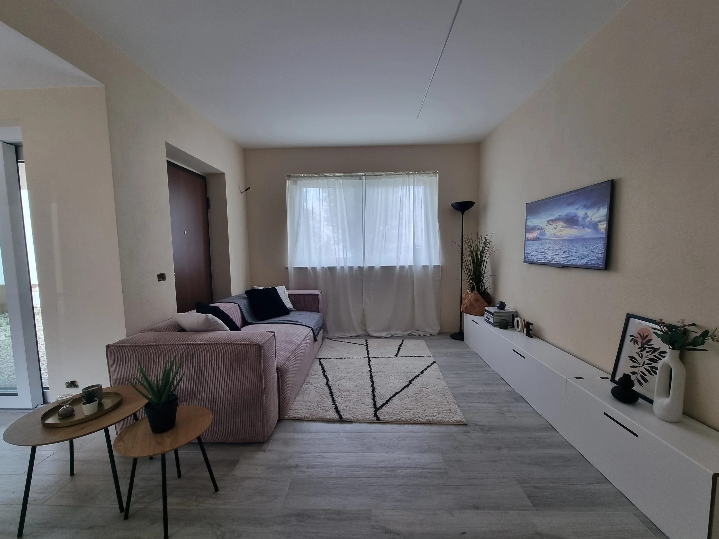 Soggiorno moderno con divano rosa, due tavolini di legno, tv a parete, tende bianche, tappeto geometrico, piante e decorazioni.