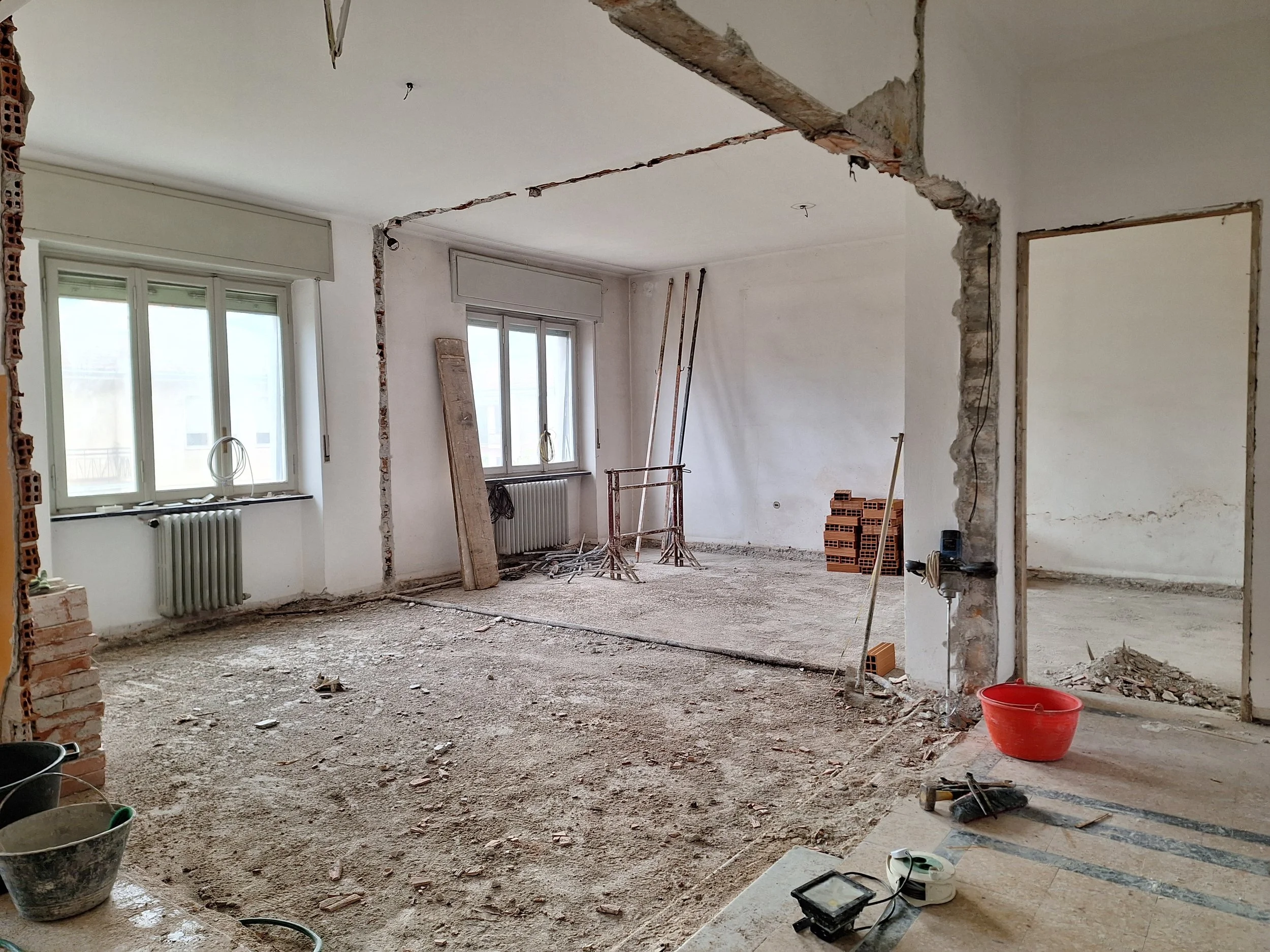 Interno di un appartamento in fase di ristrutturazione, con pavimento in asfalto e pareti in fase di demolizione, finestre sulle pareti laterali, alcuni attrezzi e materiali da costruzione sparsi.