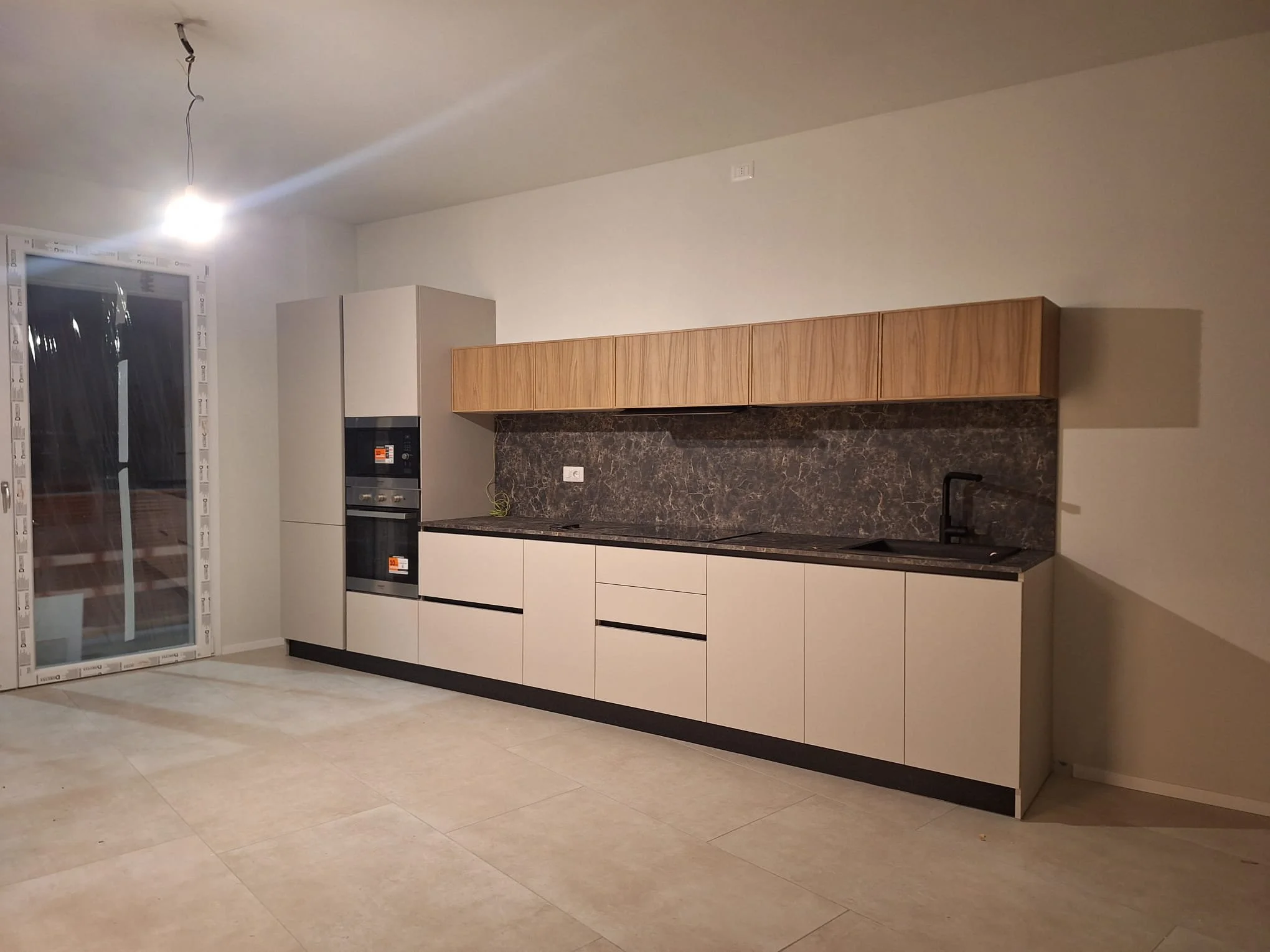 Cucina moderna con mobili bianchi e pensile in legno, piano e schermo in marmo scuro, lavandino nero e finestra.