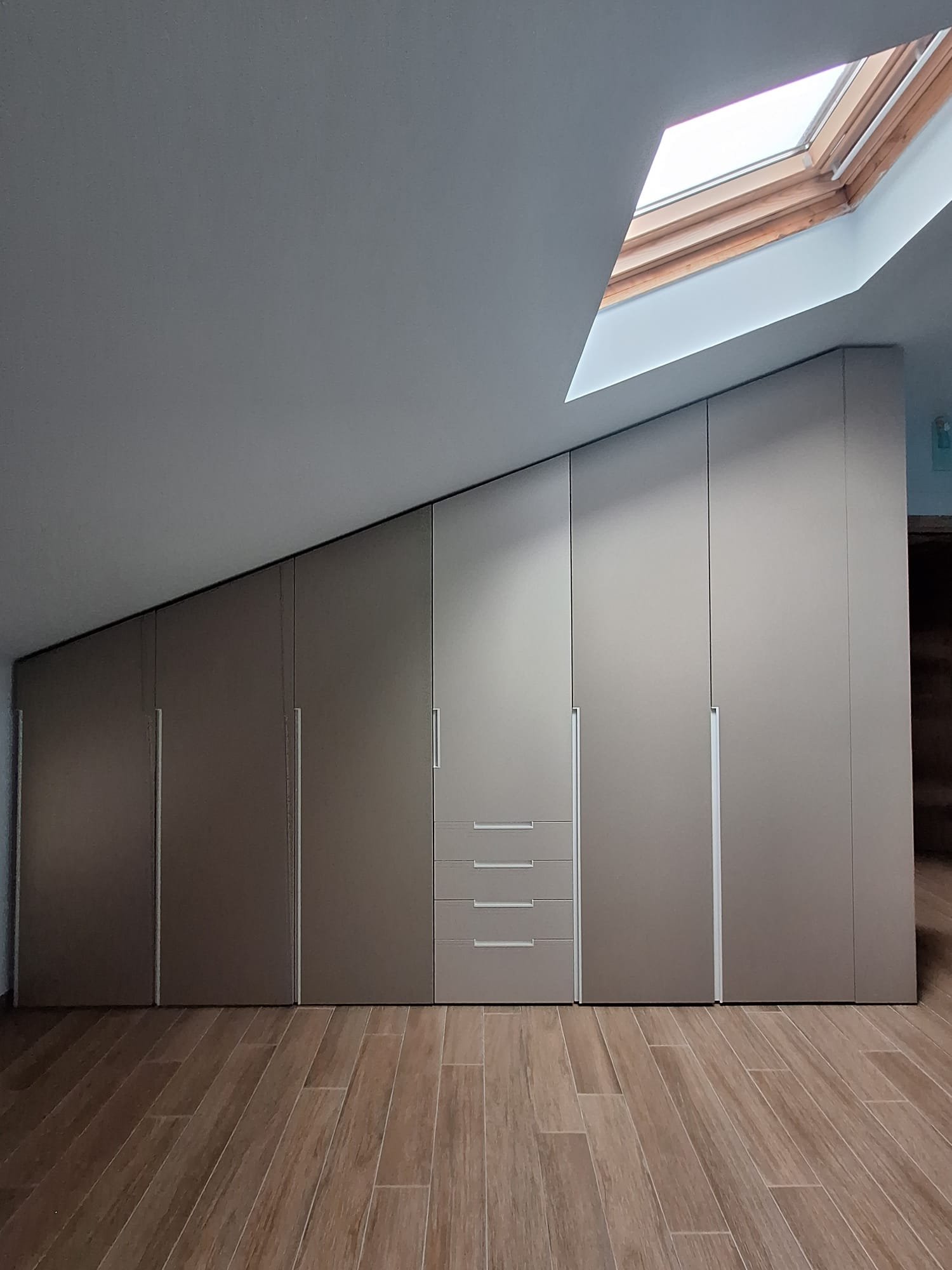 Armadio a muro in tonalità grigia con maniglie, sotto una finestra a tetto in legno, in una stanza con pavimento di legno.