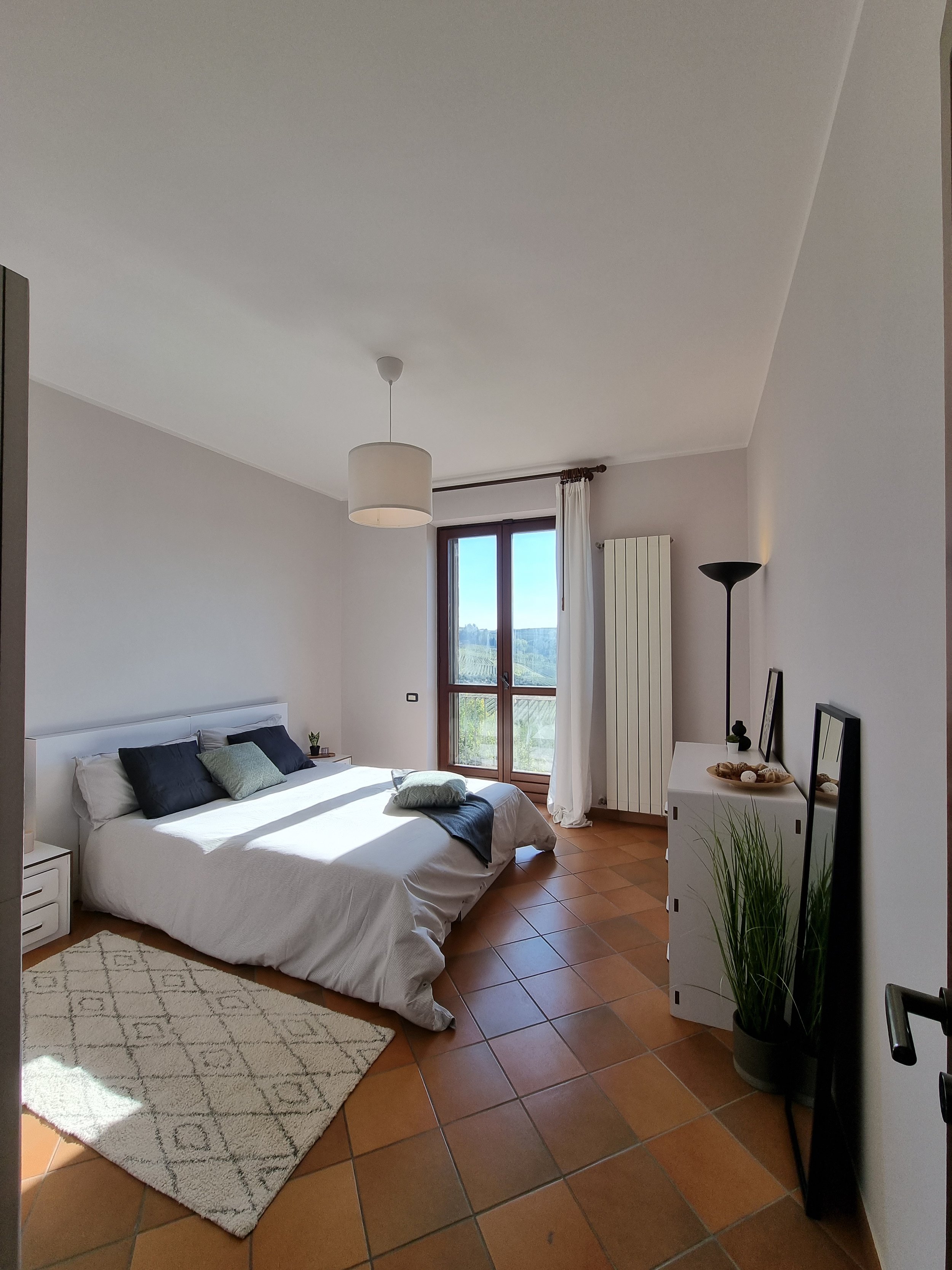 Camera da letto con letto matrimoniale, due cuscini scuri e uno chiaro, tappeto beige e pianta. Finestra con tende bianche e vista sul paesaggio, lampada da terra e uno specchio nero a terra.