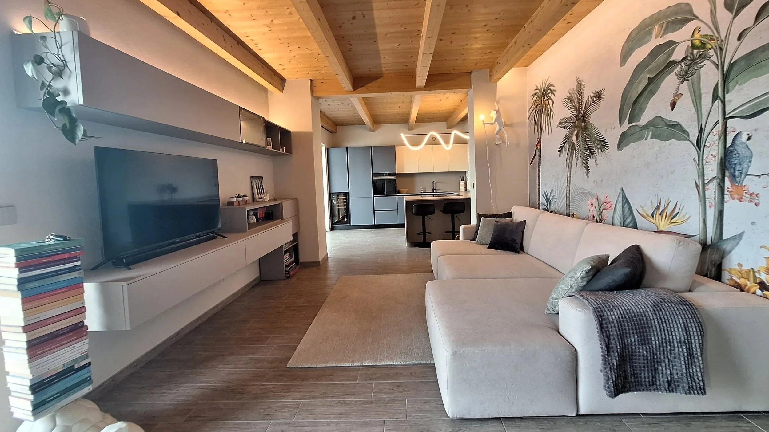 Interno di un soggiorno con divano beige, parete con wallpaper di piante e uccelli, televisore su mobile bianco, cucina moderna sullo sfondo, soffitto in legno a vista.