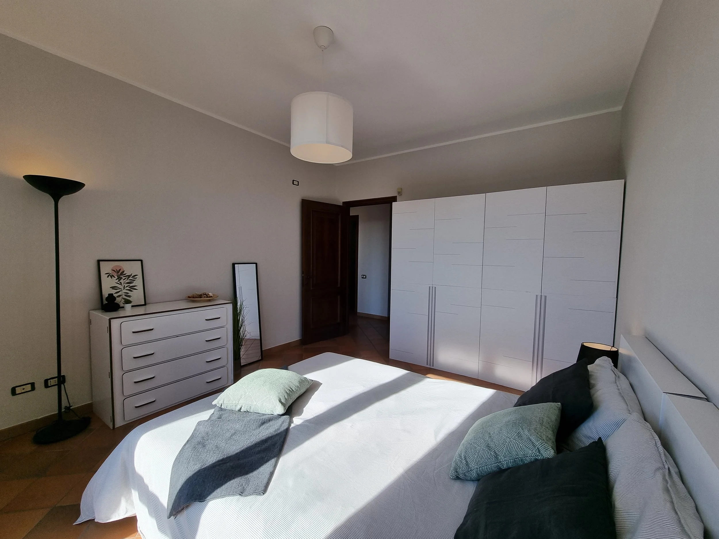 Camera da letto arredata con un letto, cuscini, una grande armadio bianca, una lampada da terra nera, un quadro e uno specchio.