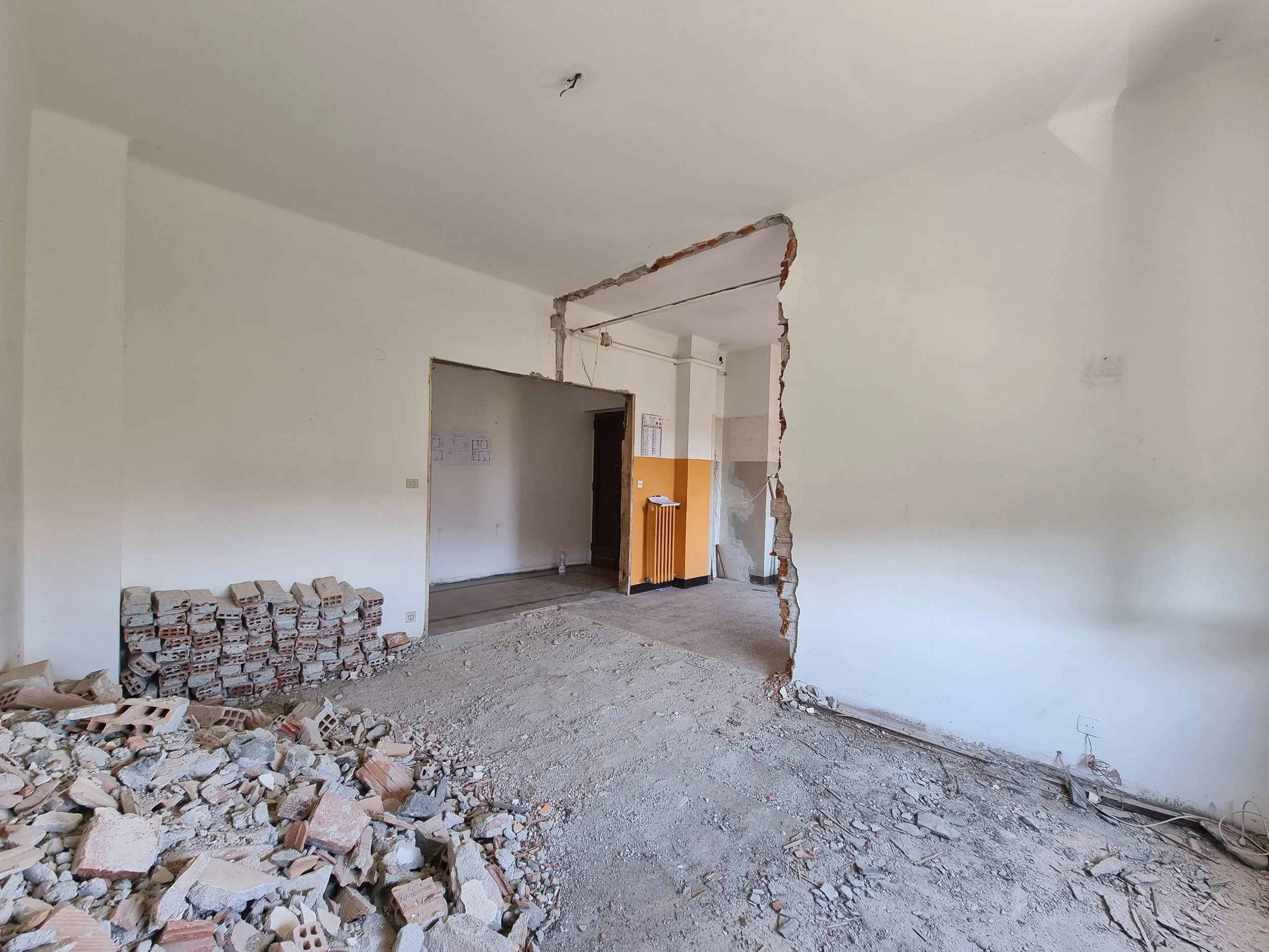 Interno di un appartamento in fase di ristrutturazione, con pareti in fase di demolizione, pile di mattoni, e cavi elettrici sospesi.