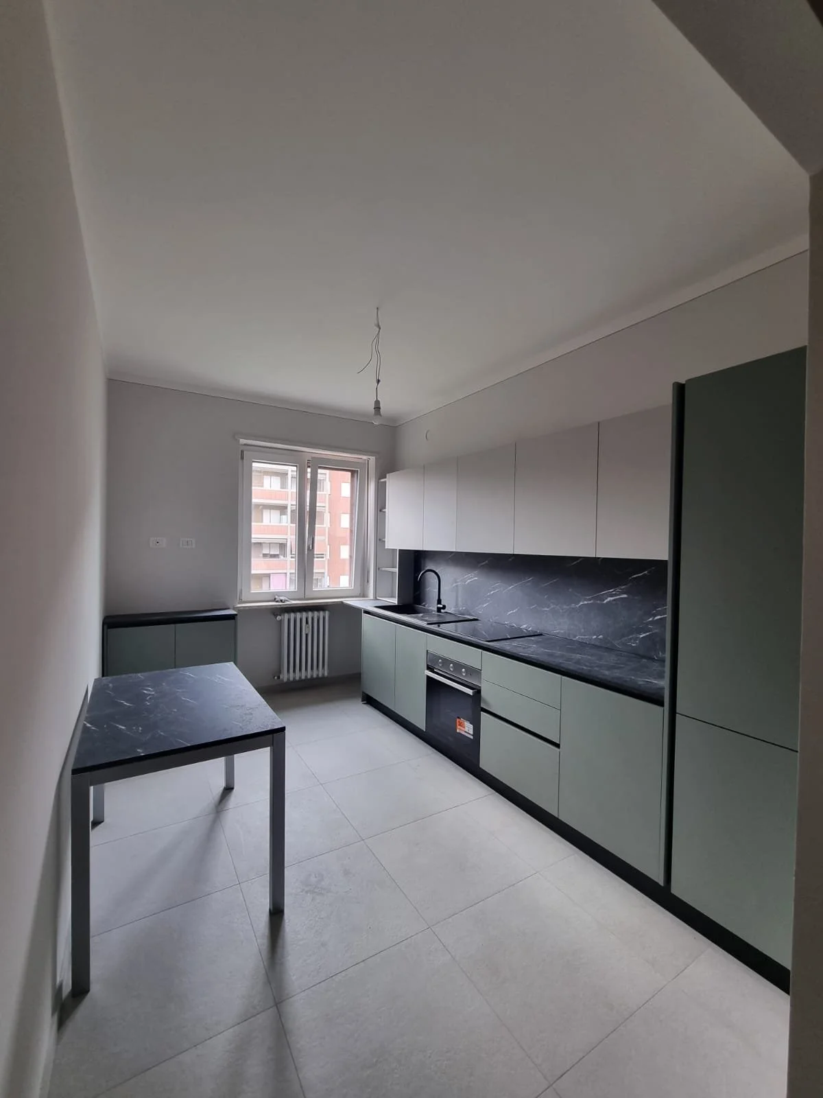 Cucina moderna con ante verdi, piano di lavoro in marmo nero, lavello nero e forno integrato. Finestra con vista su edificio residenziale e radiatore sotto la finestra.