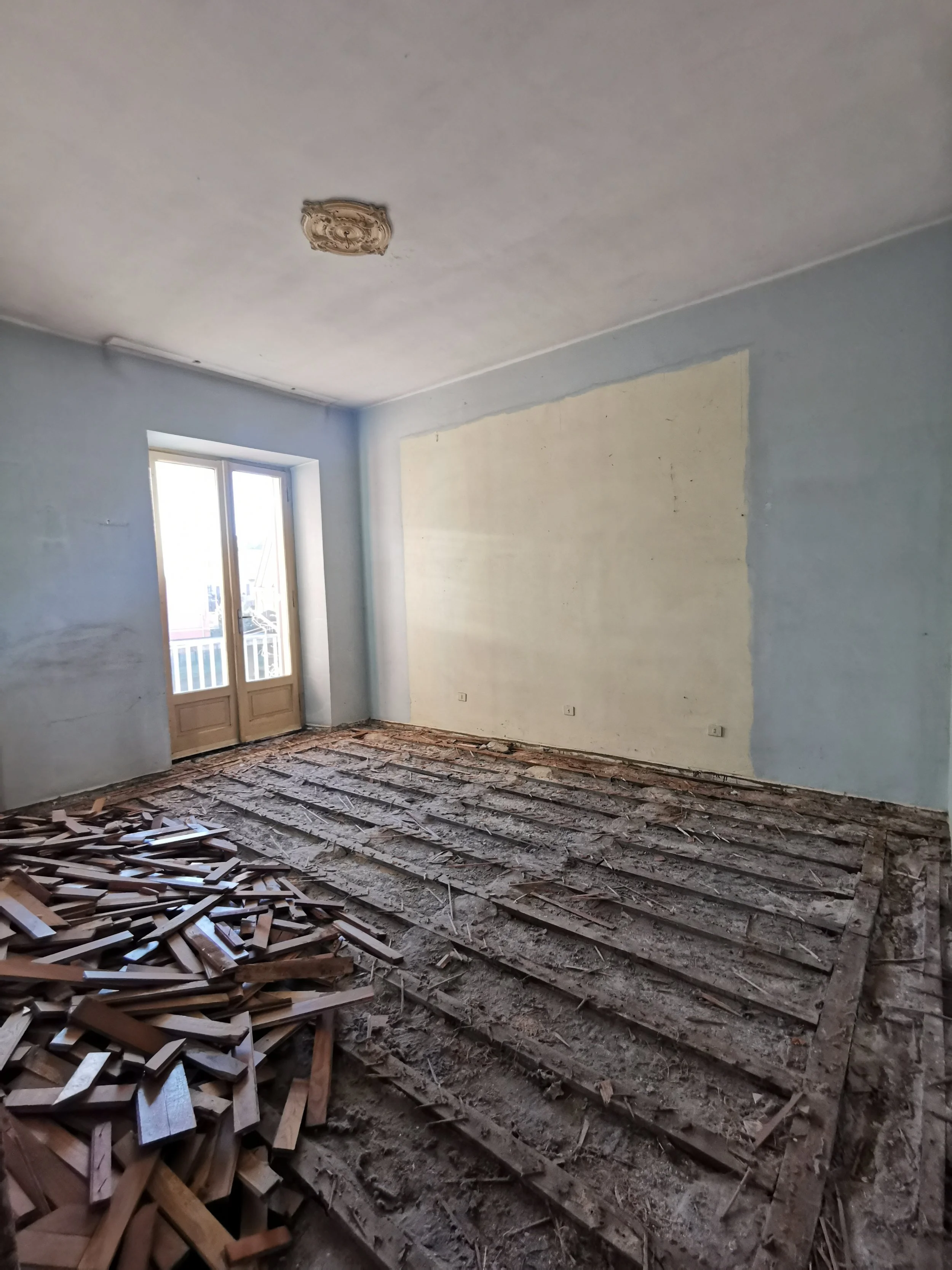Stanza in ristrutturazione con pavimento rimosso, pareti dipinte di azzurro, porta finestra con balcone, senza mobili, con immondizia e pezzi di pavimento sul pavimento.