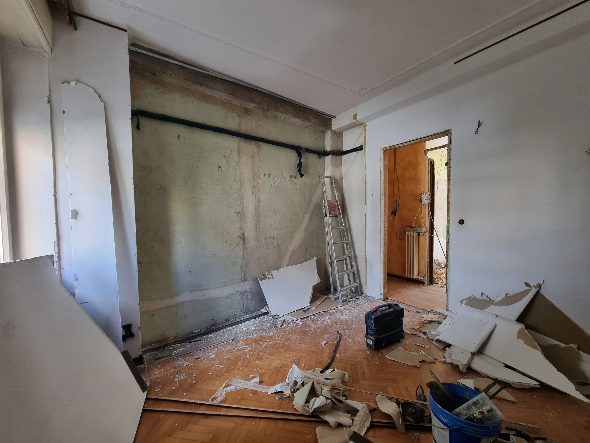 Interno di una stanza in ristrutturazione con muri danneggiati, attrezzi e detriti sul pavimento, scala e apertura verso altra stanza con radiator.