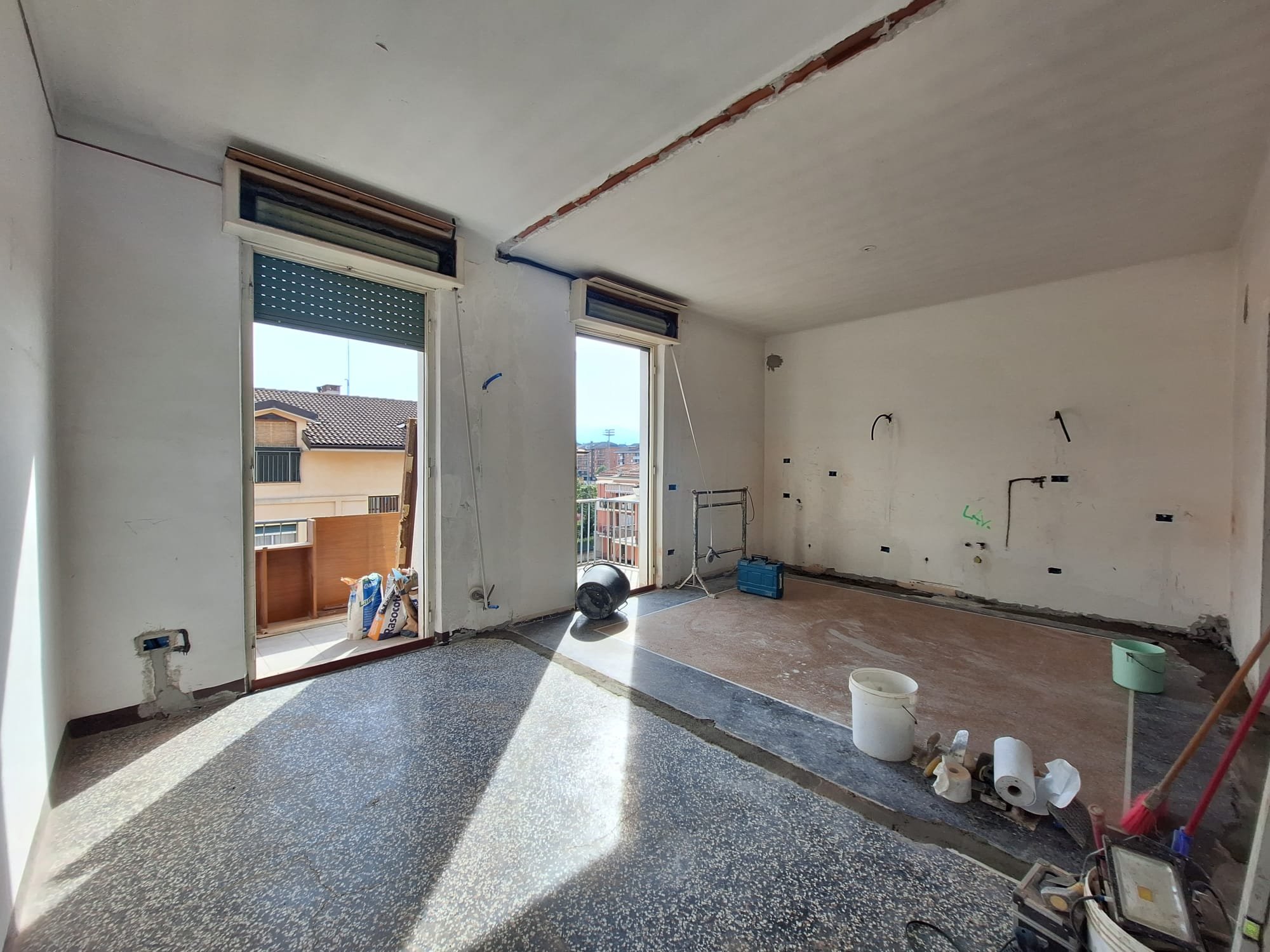 Interno di una stanza in fase di ristrutturazione con porte-finestre che portano a un balcone, pavimento in granito, pareti vuote con alcuni allacciamenti e attrezzi da lavoro.