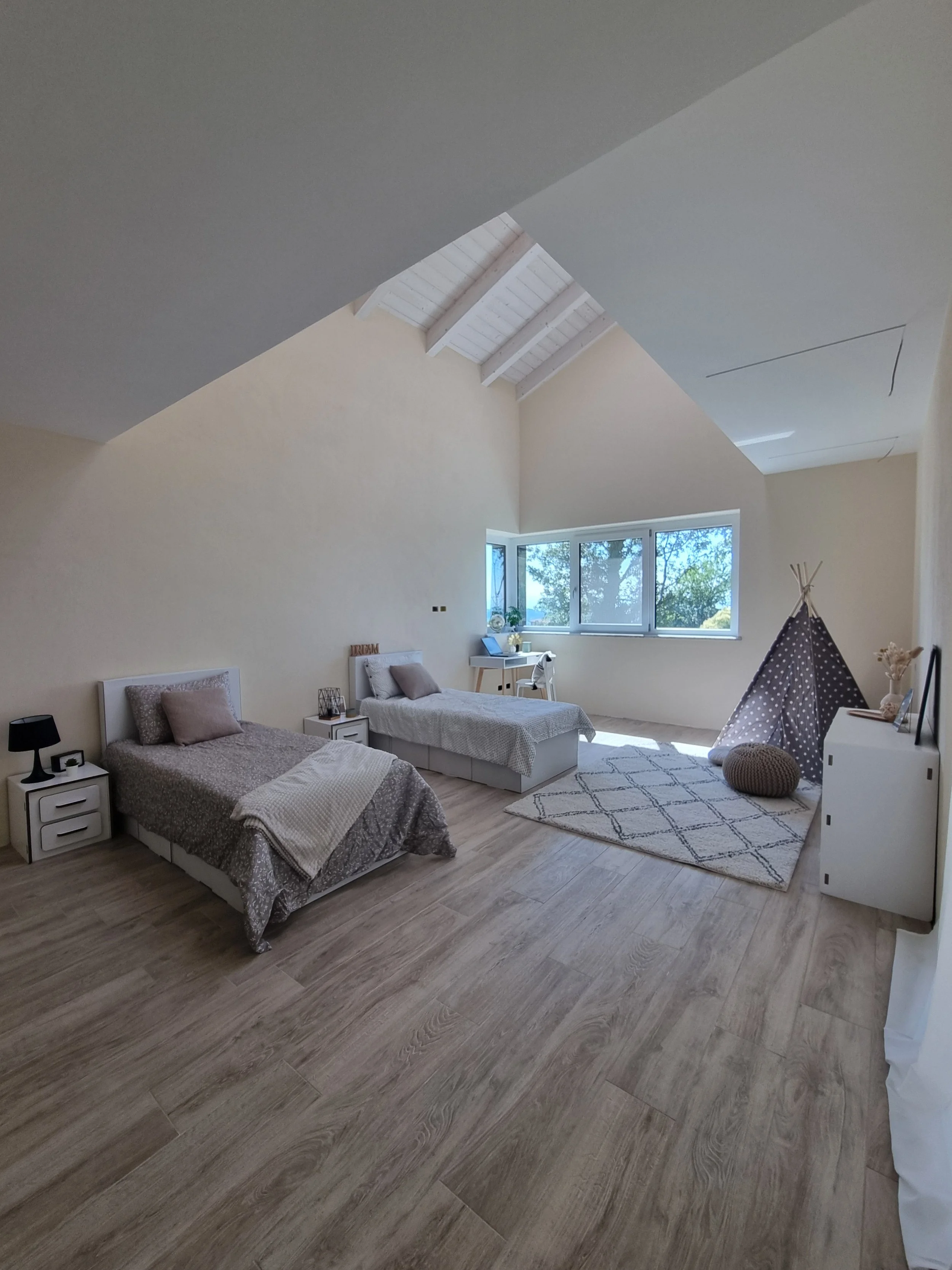 Camera da letto in stile minimalista con due letti singoli, tappeto, una tenda da gioco a forma di teepee, grande finestra e soffitto alto con travi di legno bianco.