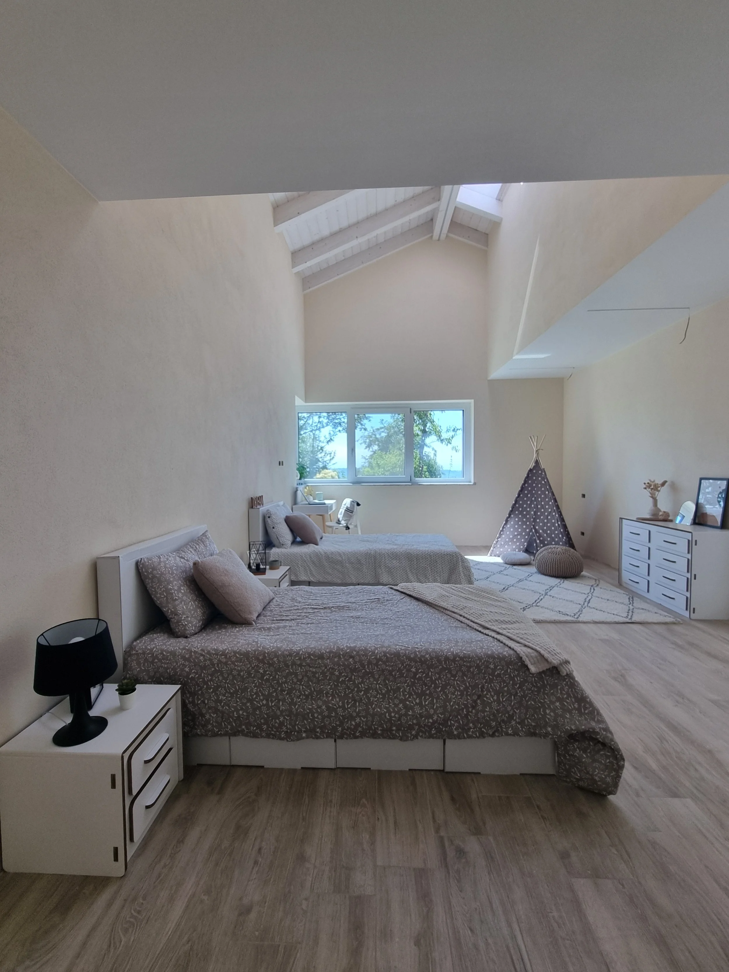 Camera da letto spaziosa con due letti, grande finestra con vista sul verde, zona giochi con tenda a teepee e pouf, armadio bianco e pavimento in legno chiaro.