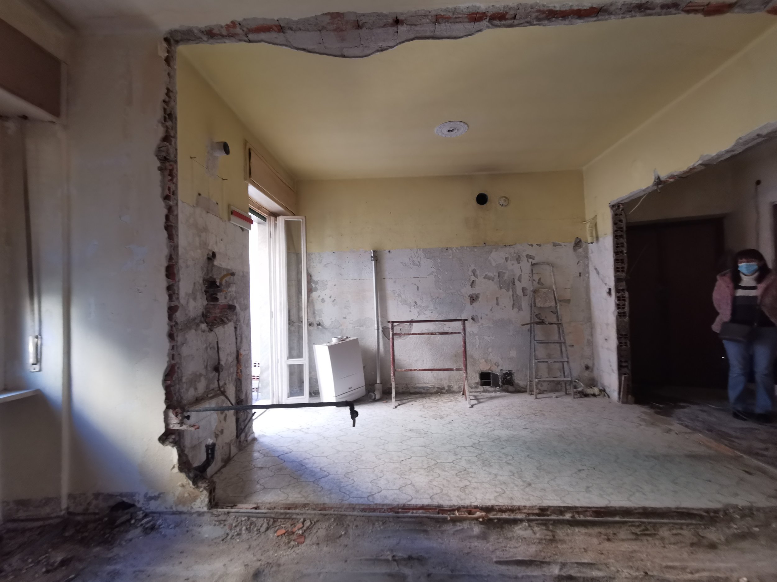 Interno di una stanza in ristrutturazione con pareti rovesciate, scala, scaffale e finestra aperta, con una persona con mascherina che entra dalla destra.