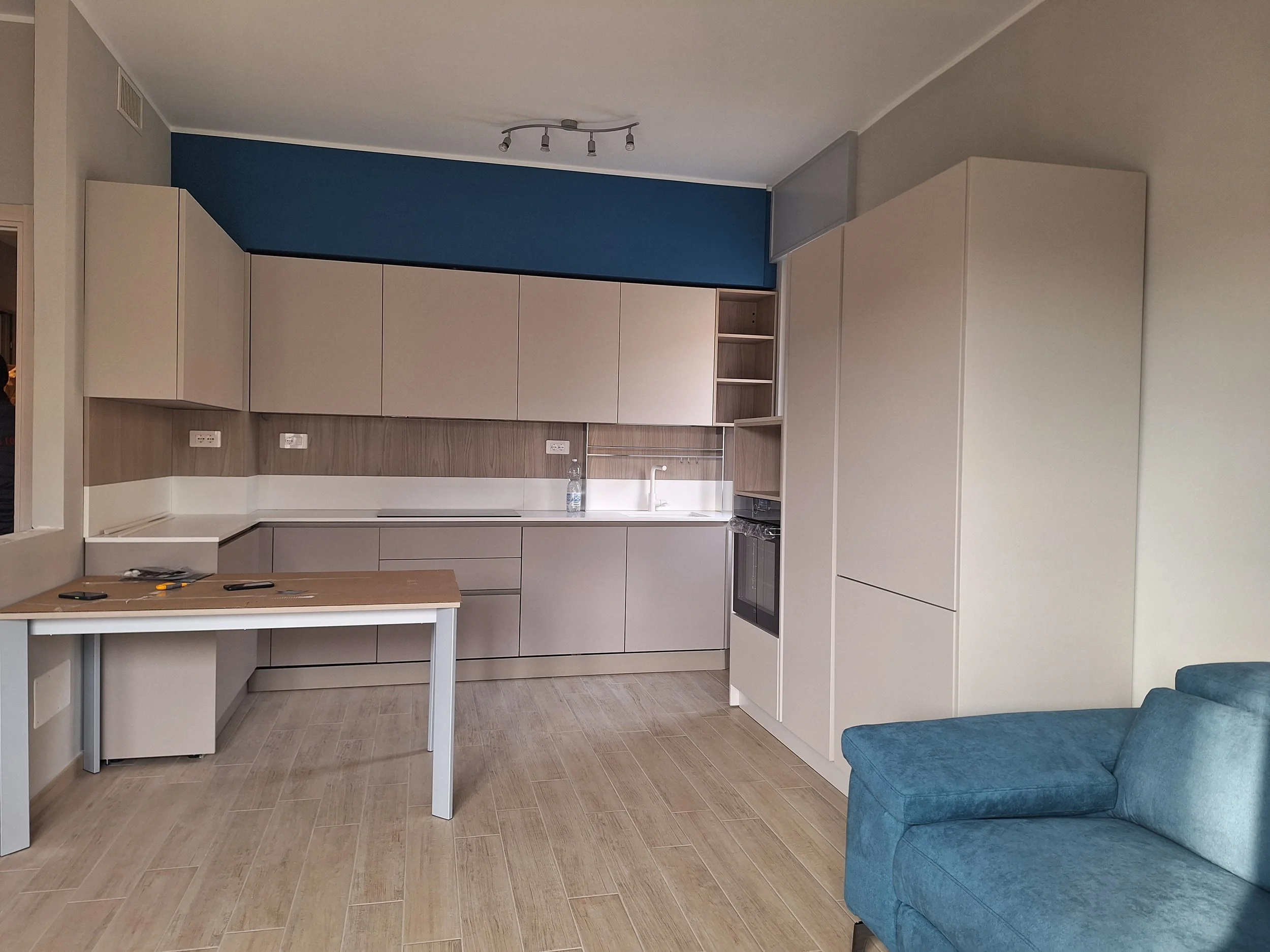 Cucina moderna con armadi beige, piano di lavoro bianco, parete di legno, e angolo con scaffali aperti davanti a una parete blu. In primo piano, tavolo di legno con strumenti e una bottiglia di acqua, e divano azzurro a fianco.