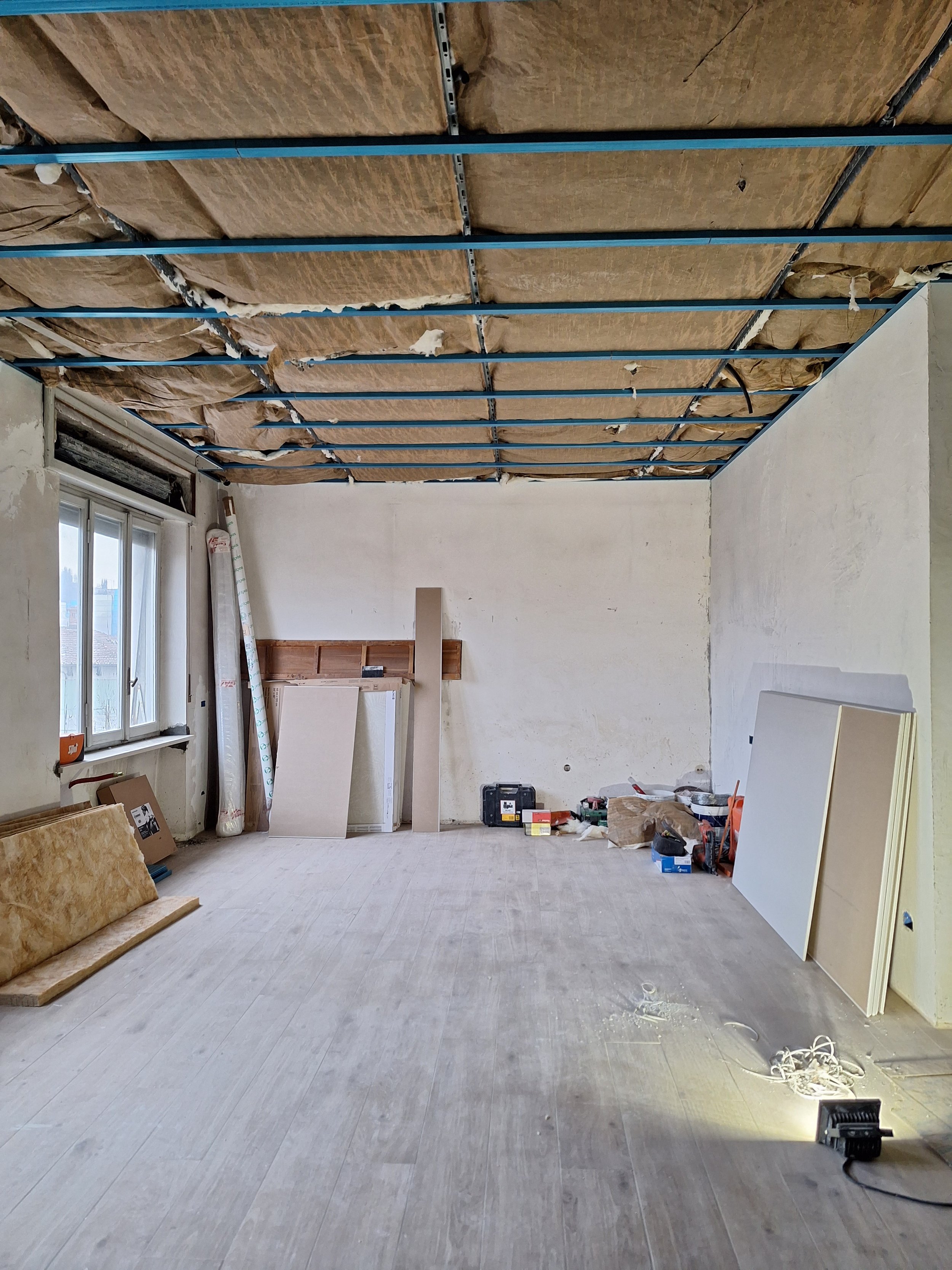 Stanza in ristrutturazione con pareti dipinte di bianco, finestrella, e soffitto senza finitura con assi e supporti visibili, materiali e attrezzi di lavoro sparsi su pavimento in legno.