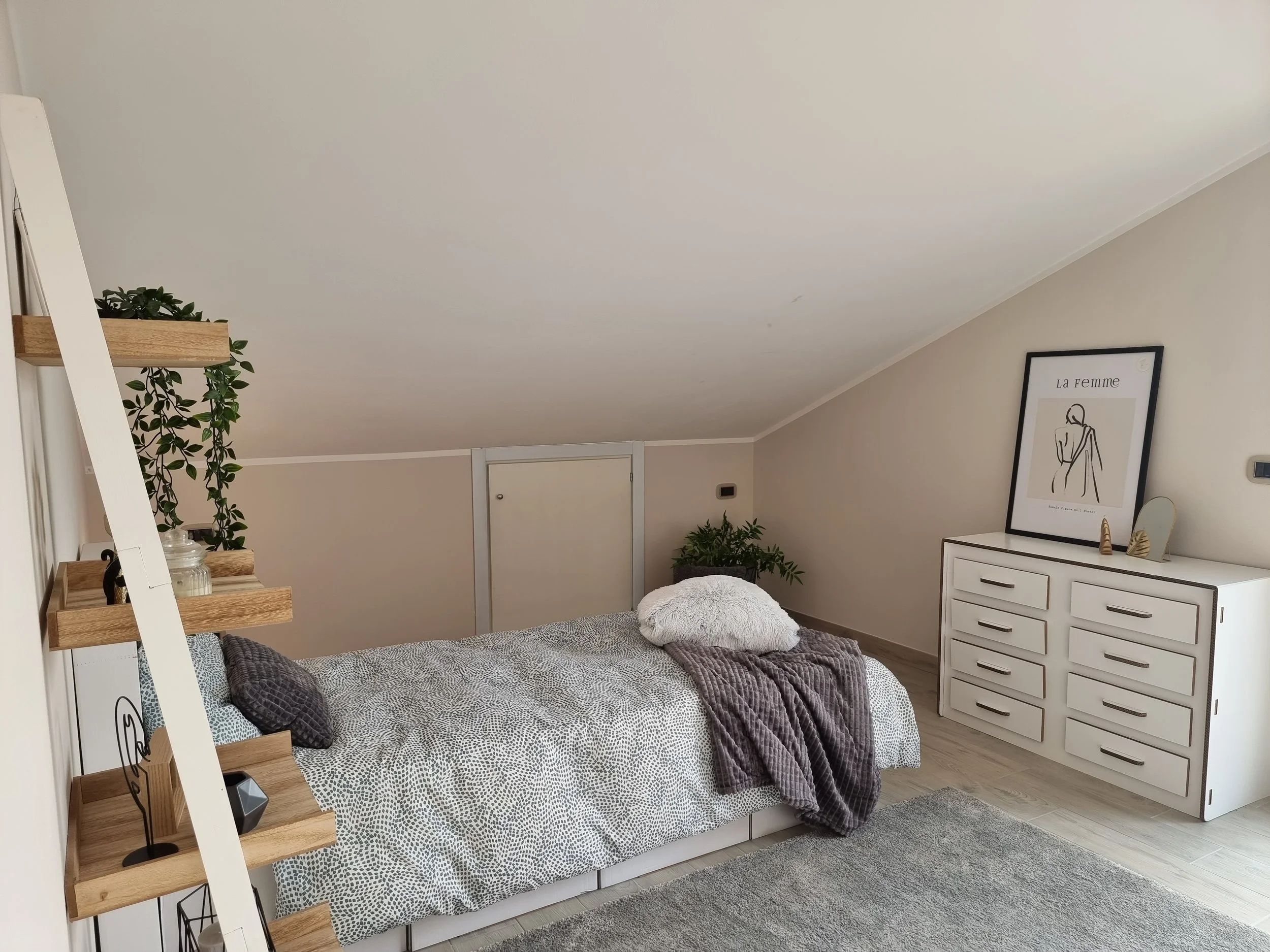 Una camera da letto con un letto matrimoniale, coperte grigie e cuscini, scaffali in legno con decorazioni e piante, una cassettiera bianca con un quadro e oggetti decorativi, pareti bianche, pavimento in legno e un tappeto grigio.