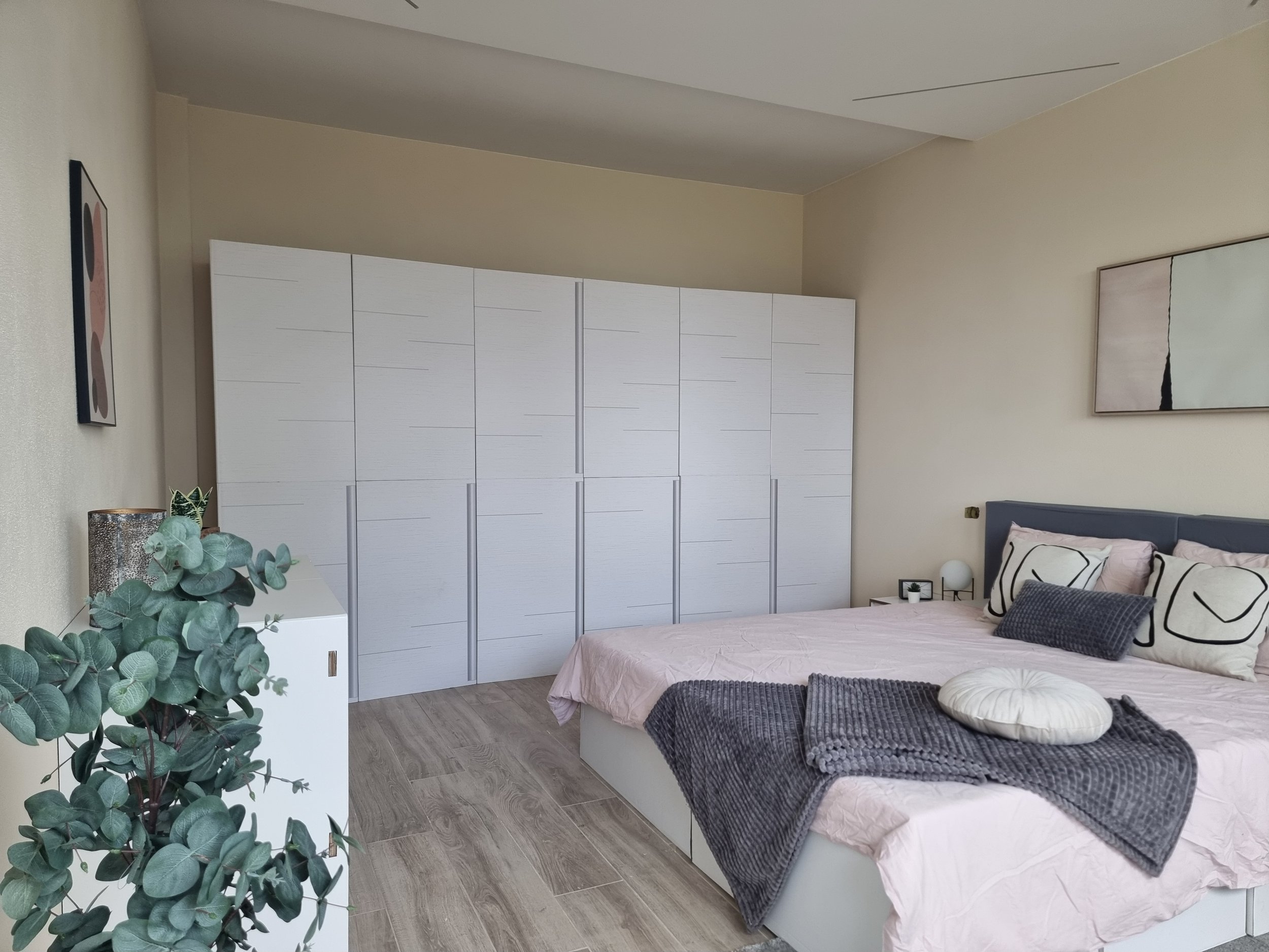 Una camera da letto con pareti beige, un letto con cuscini e lenzuola rosa, un grande armadio bianco con ante impiallacciate e decorazioni minimaliste, un comodino con decorazioni e una pianta in primo piano a sinistra.