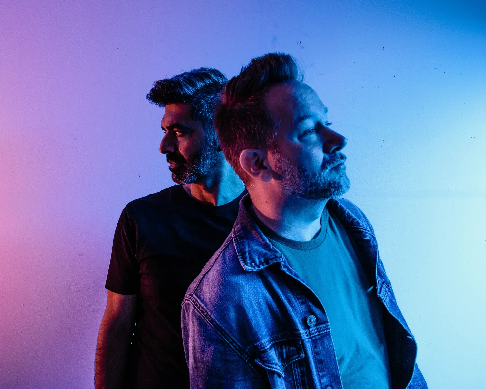 Two men standing back to back against a gradient background of purple, pink, and blue.