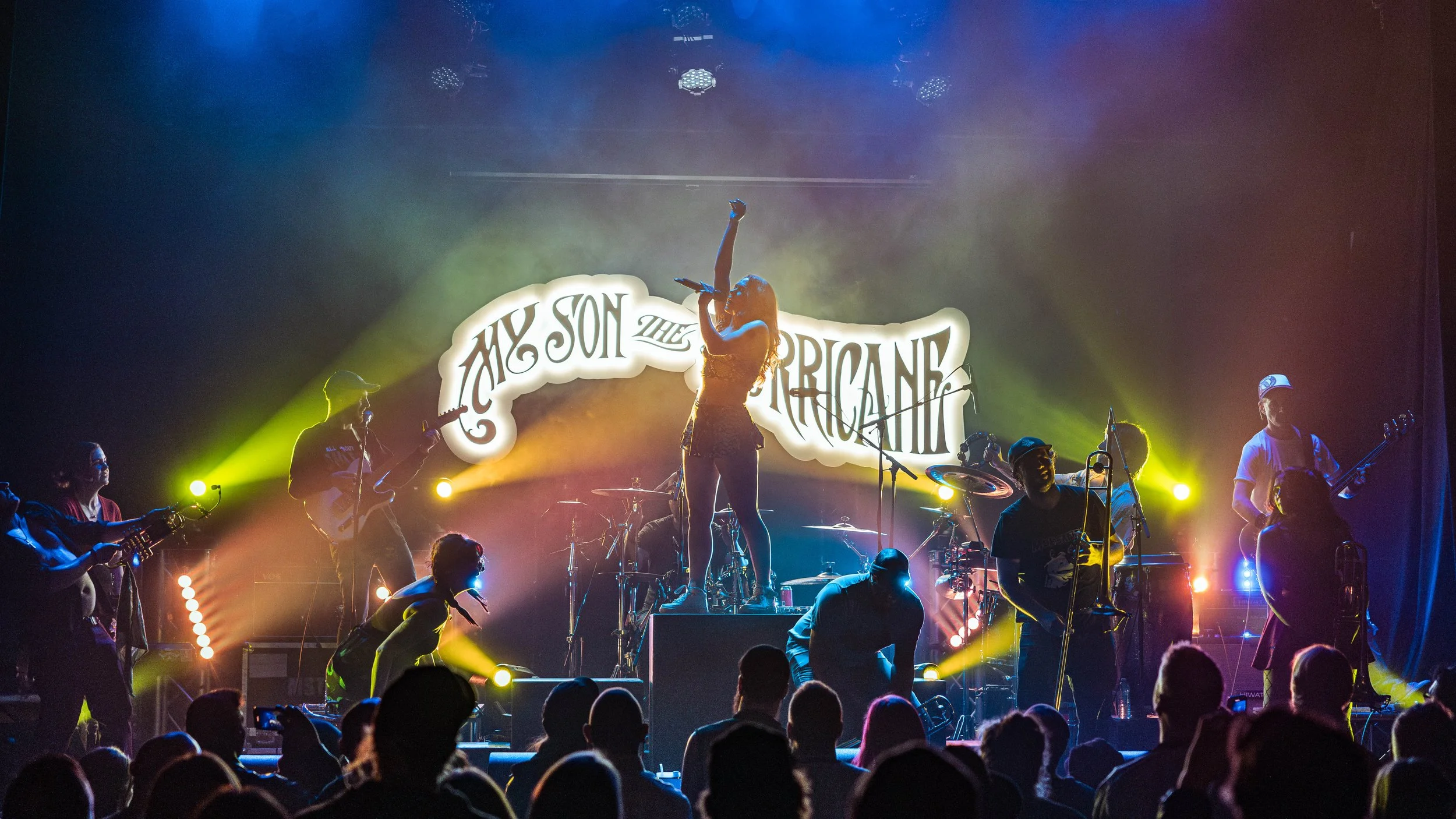 A live concert scene featuring a female singer standing on a drum platform with her hand raised, performing on stage with a large illuminated sign behind her reading "Amy Son the Tropical." Several musicians are playing instruments around her, with colorful stage lighting and an audience in silhouette watching.