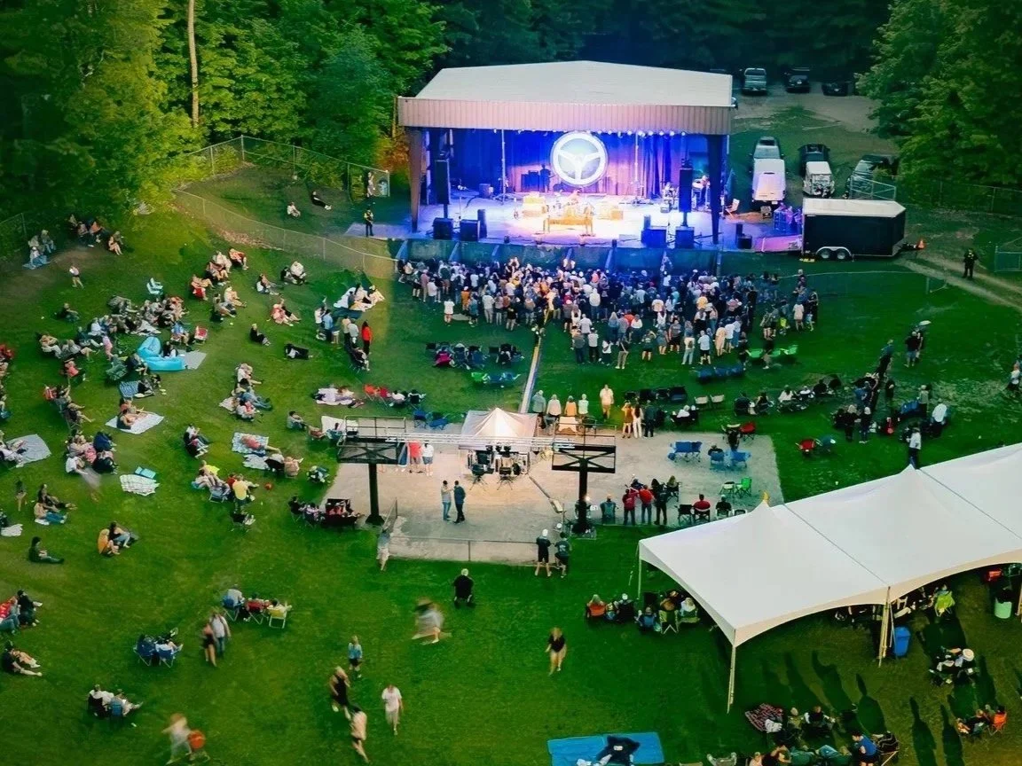 Outdoor concert in a park with a stage, audience seated on the grass, some on blankets, and some under a canopy tent.