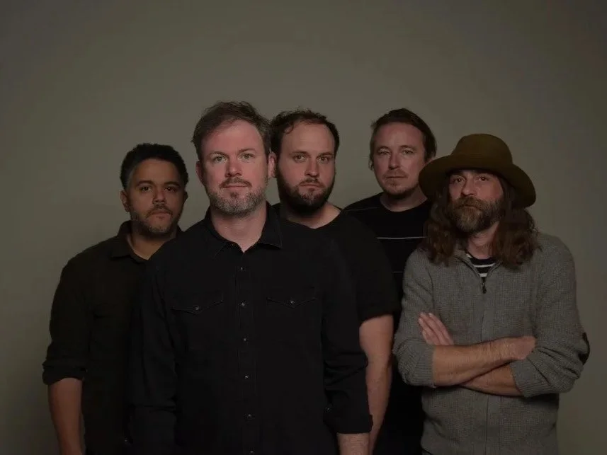 A group of five men standing against a plain background, dressed in dark casual clothing, with one wearing a hat.