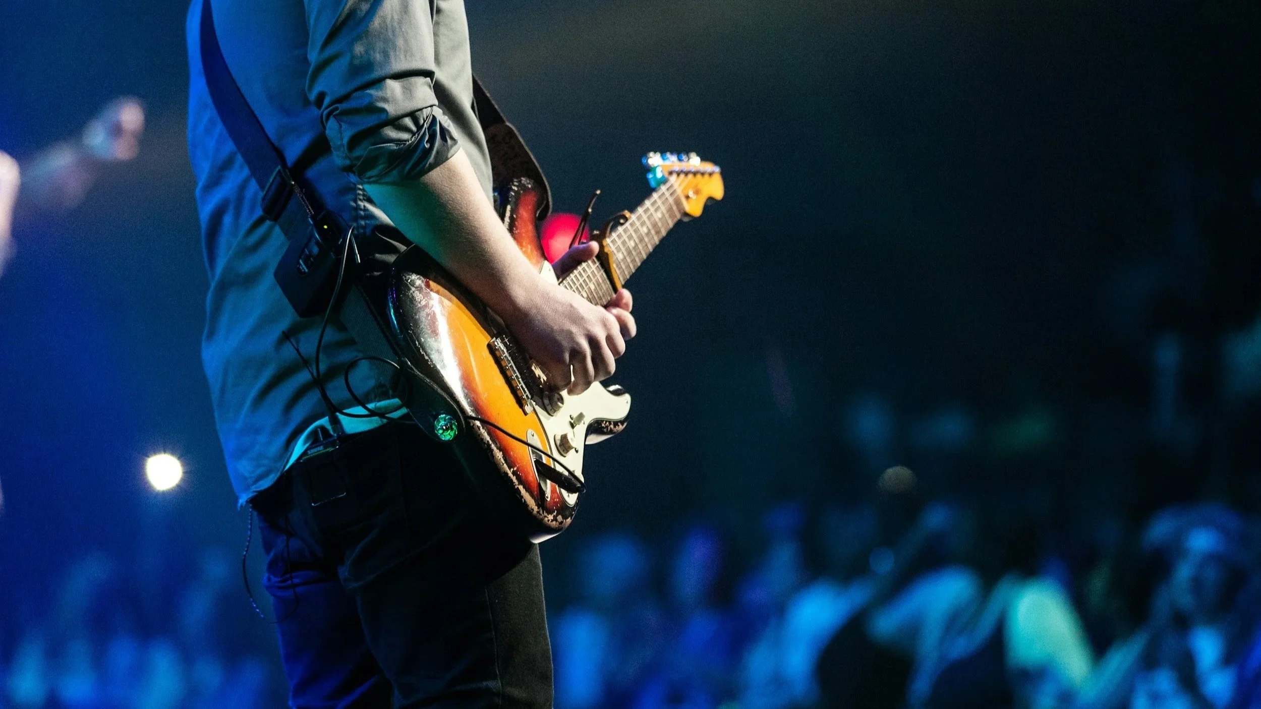 Person playing an electric guitar on stage during a concert, with an audience in the background.