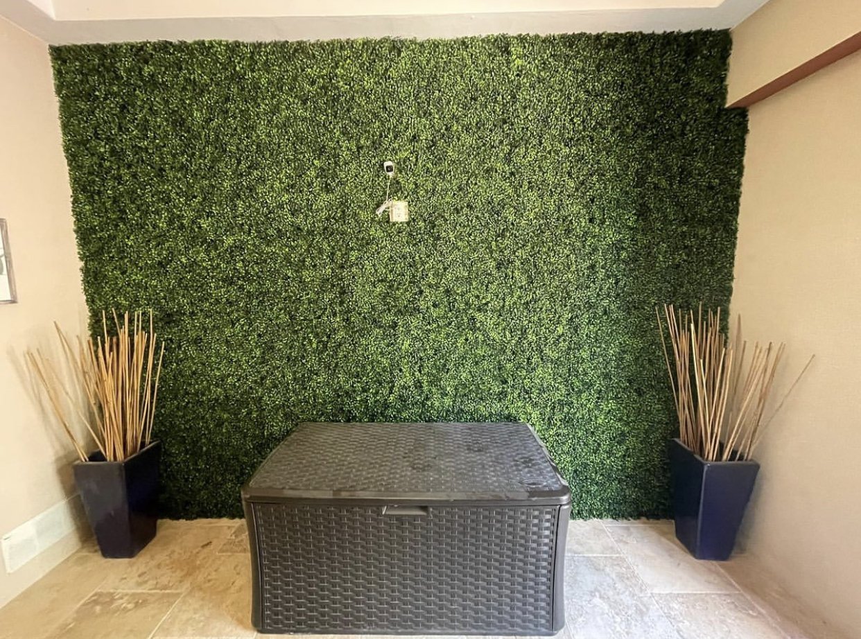 Indoor space with a green hedge wall, a black storage box, and two tall black planters with wooden sticks on either side.