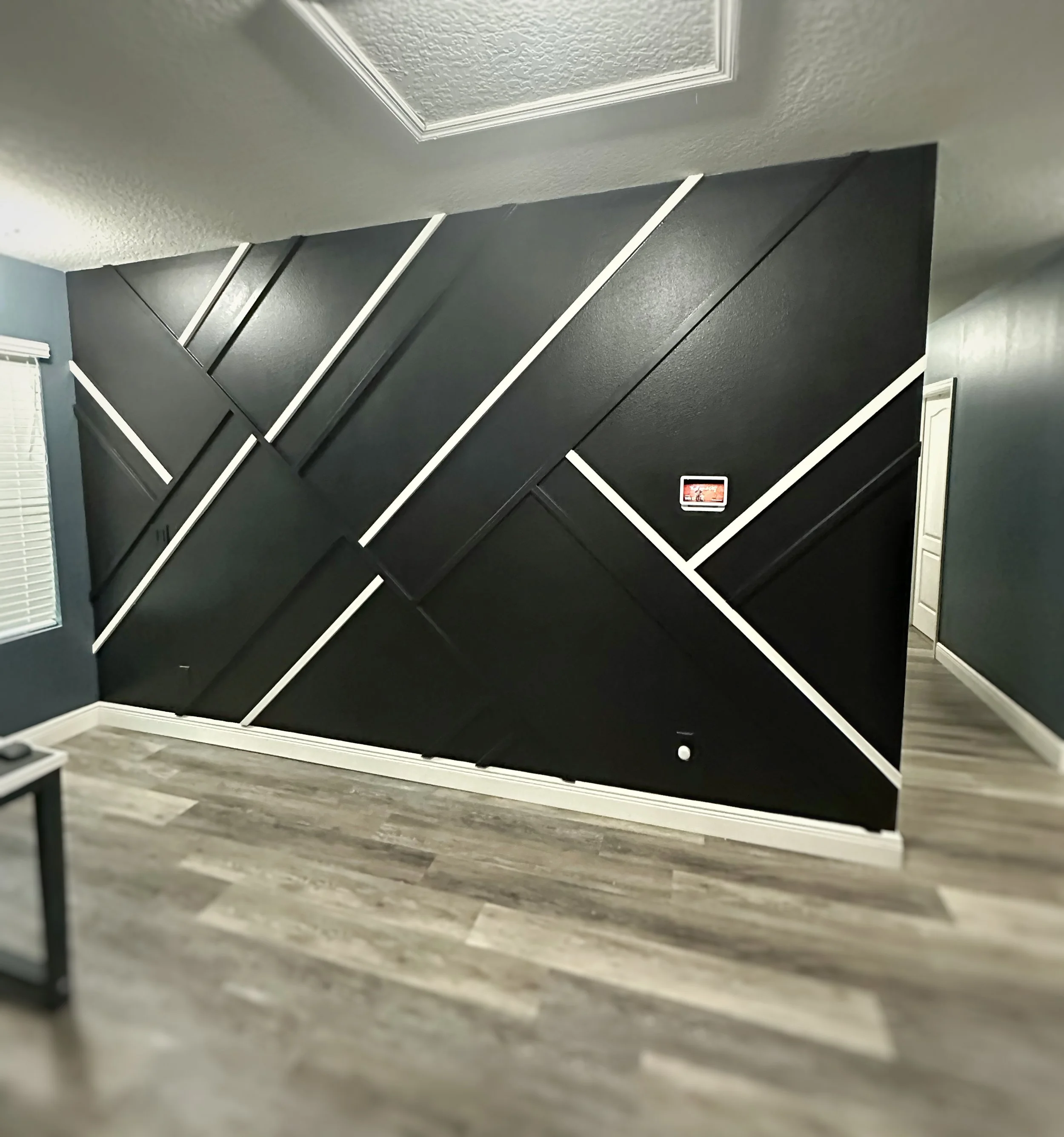 Interior of a room with a black geometric accent wall, light wood flooring, and a ceiling vent.