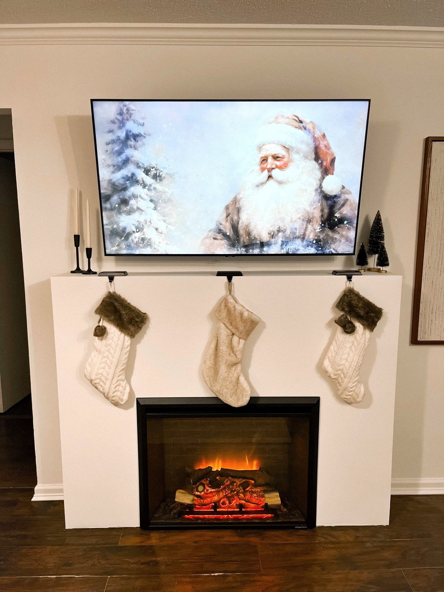 A living room fireplace mantel decorated for Christmas with three stockings hanging, a flat-screen TV displaying an image of Santa Claus in a snowy landscape, and small decorative Christmas trees on the side of the mantel, with an electric fireplace 