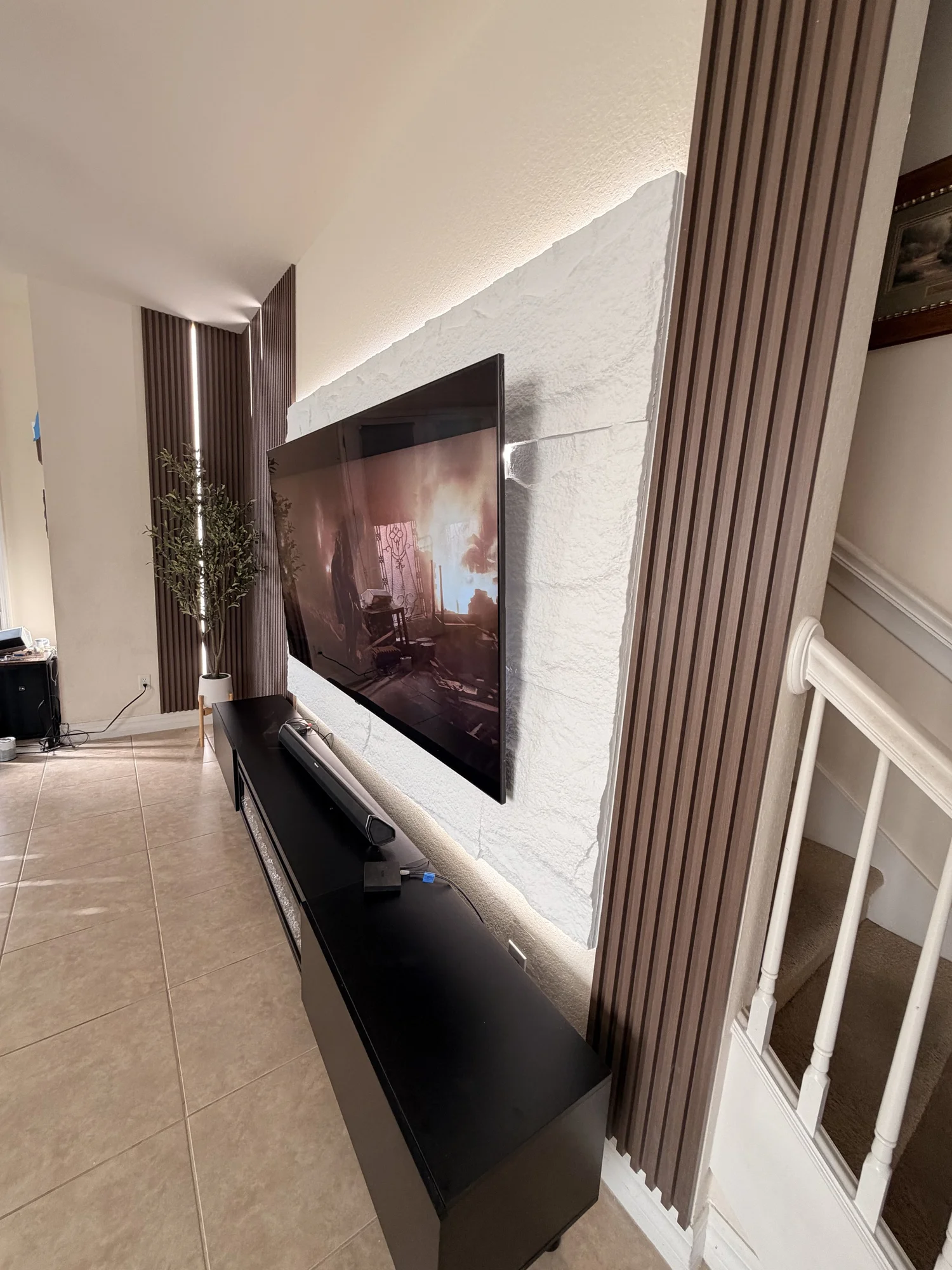 A living room corner featuring a wall-mounted flat-screen TV, a black soundbar below, a white staircase railing on the right, a tall potted plant next to brown wooden slat wall panels, and a beige tiled floor.
