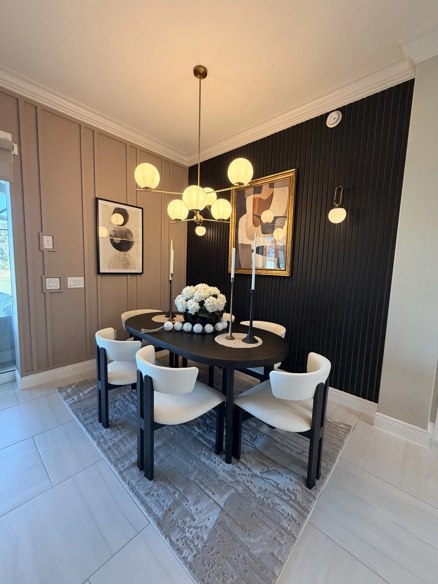 A dining room with a black oval table surrounded by six white upholstered chairs, a black and white area rug, a modern chandelier with spherical bulbs, a floral centerpiece, framed abstract artwork, black candlesticks, and black wall paneling.