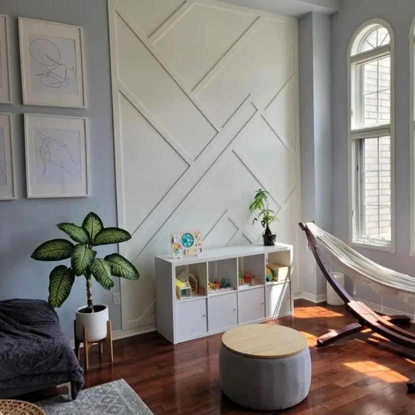Bright living room with white geometric wall design, large arched window, potted plant, white storage unit with toys, hammock chair, gray ottoman, and wooden floor.