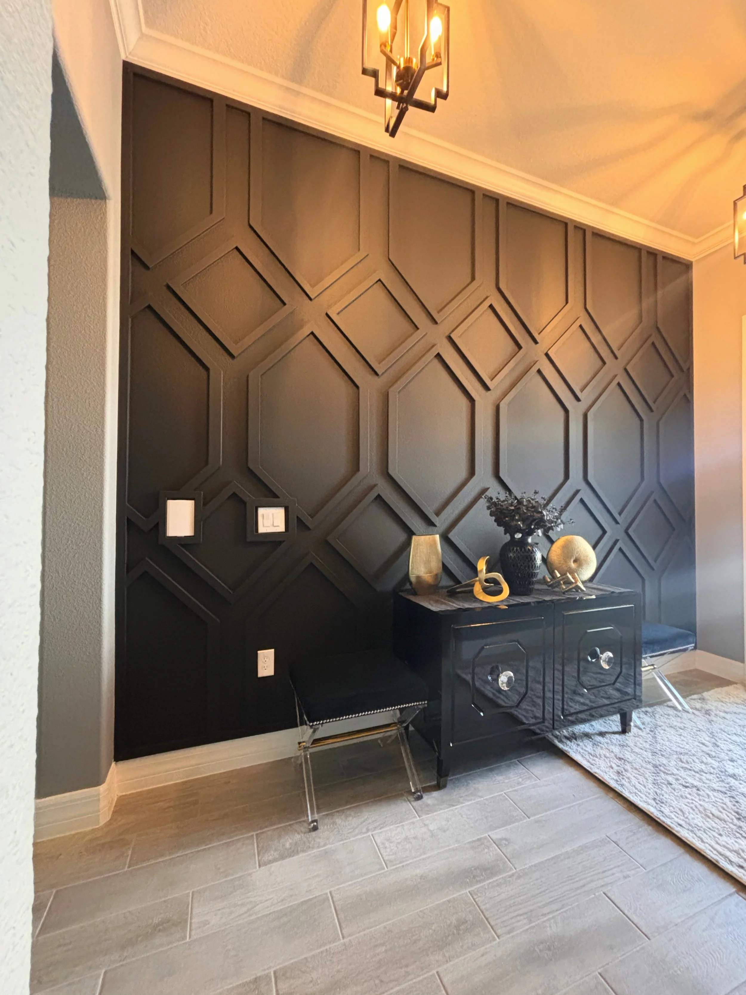 A modern interior with a black sideboard, geometric wall paneling, decorative vases, and two black chairs with glass legs, illuminated by patterned ceiling lights.